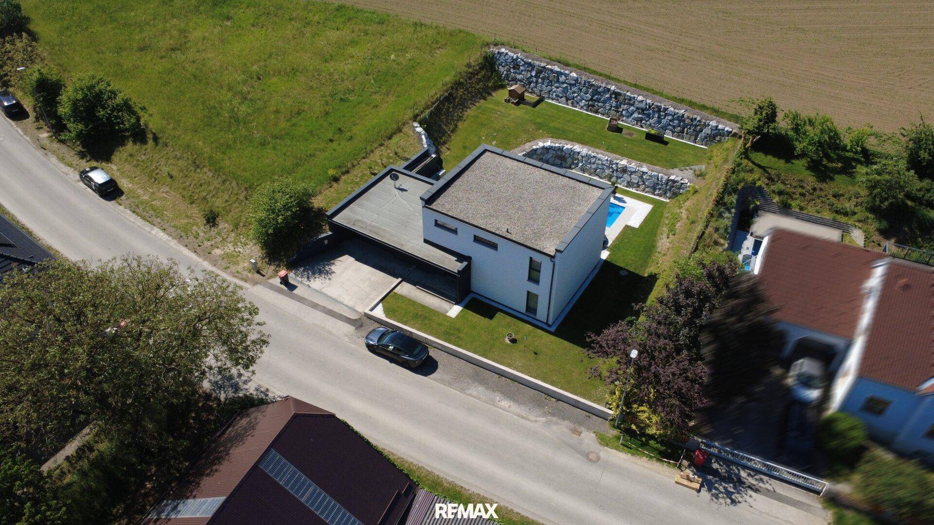 Vogelperspektive des Hauses mit Flachdach, Zufahrt und angrenzenden Feldern unter blauem Himmel.