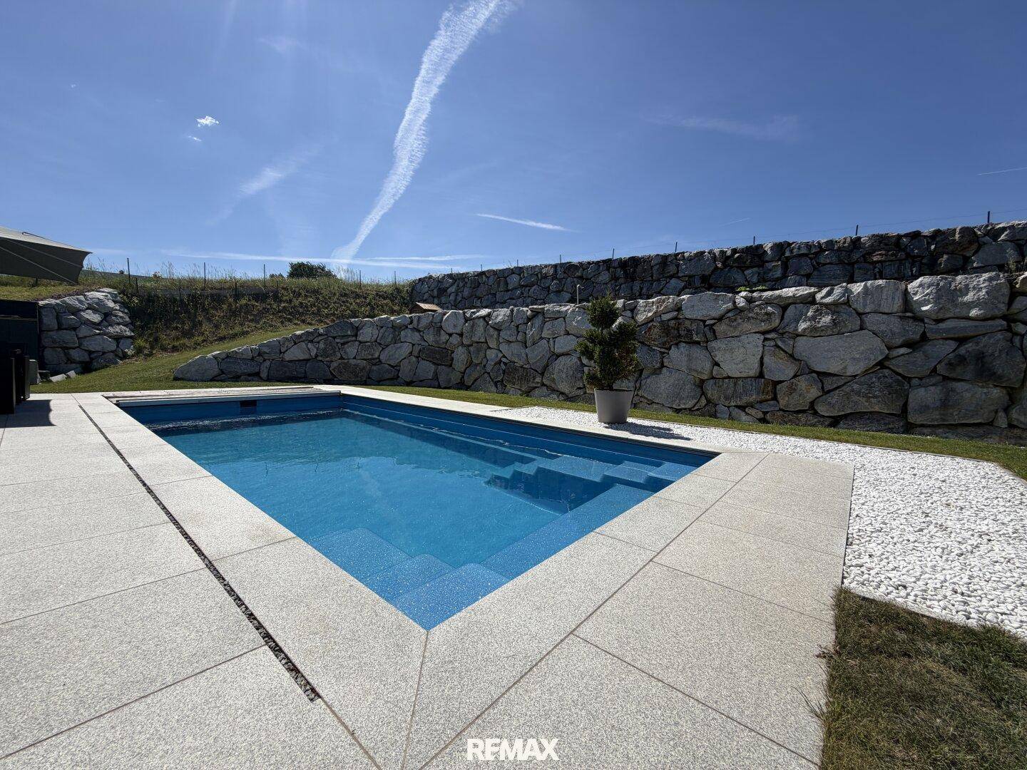 Blauer Swimmingpool mit gepflasterter Terrasse, umgeben von Rasen und einer Natursteinmauer unter blauem Himmel.