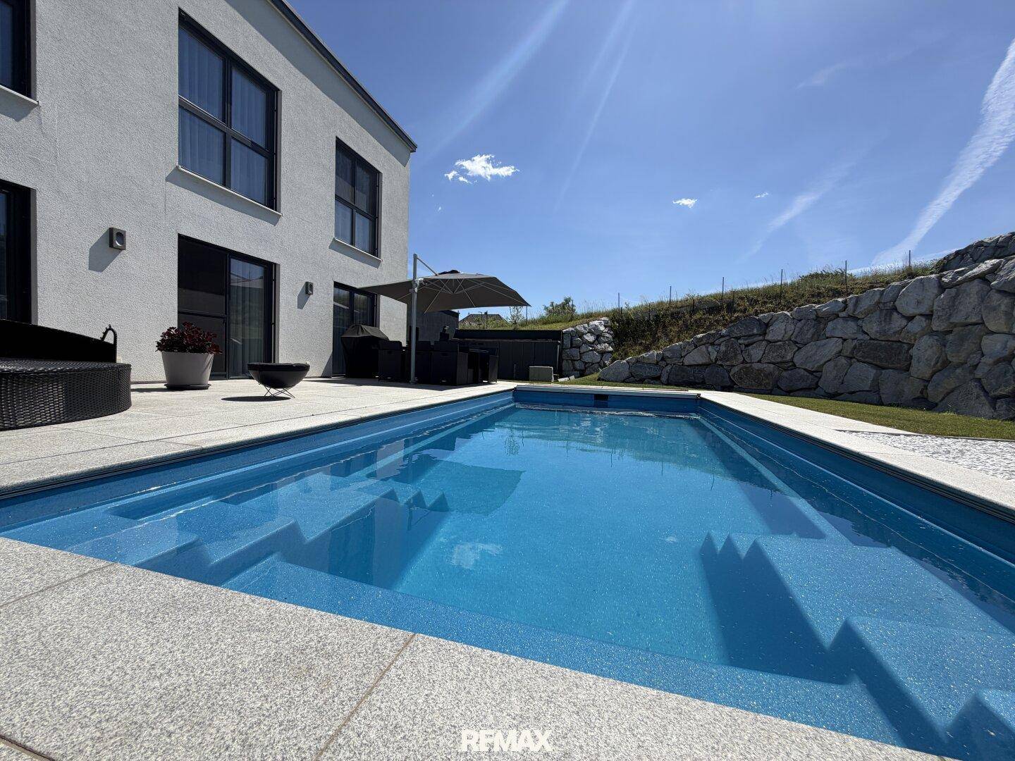 Moderner Swimmingpool mit angrenzender Terrasse und stilvollen Gartenmöbeln vor der weißen Hausfassade.