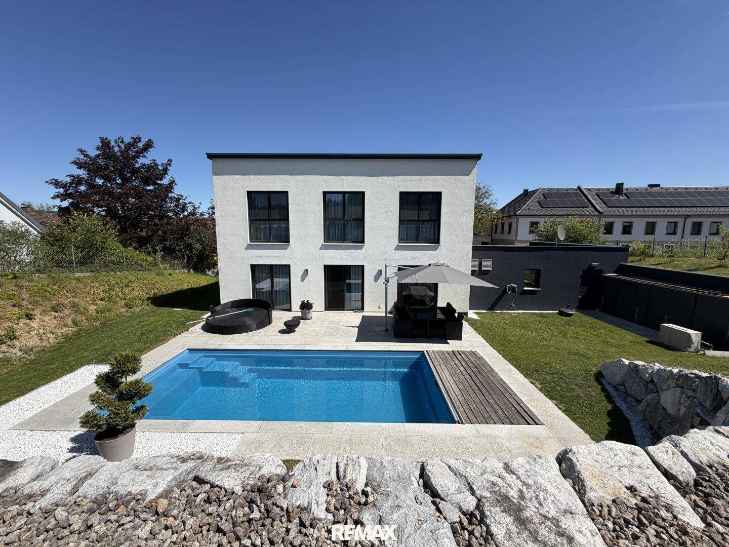 Modernes Einfamilienhaus mit Swimmingpool, Holzdeck und weitläufigem Garten unter strahlend blauem Himmel.
