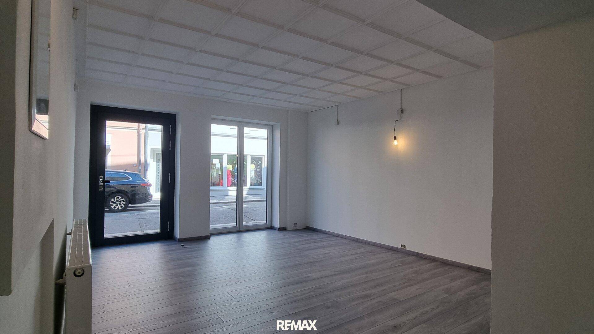 Heller, leerer Raum im Erdgeschoss mit großen Fenstern zur Straße und modernem Laminatboden.