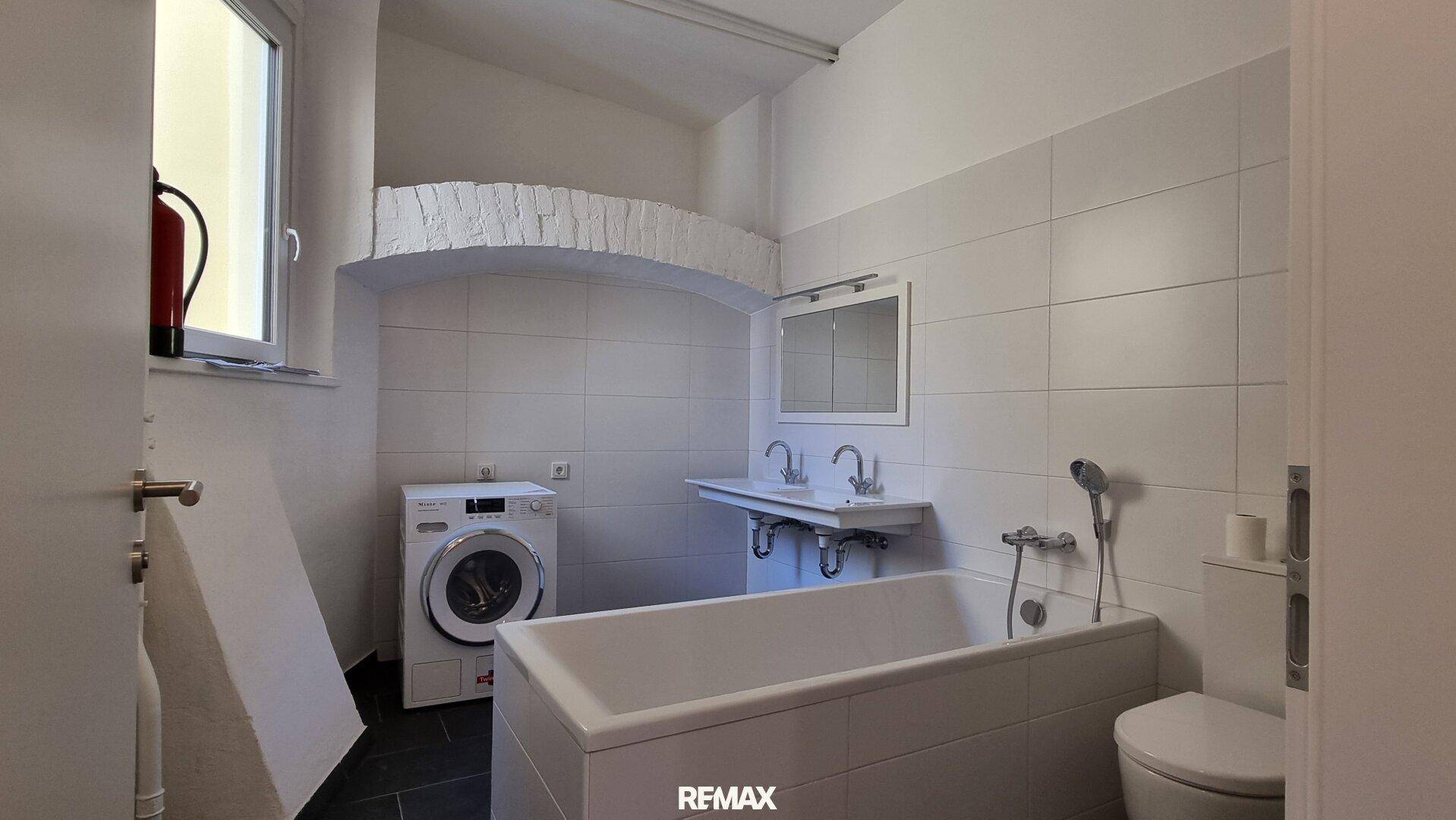 Modernes Badezimmer mit Badewanne, Doppelwaschbecken und Waschmaschine, hell gefliest.
