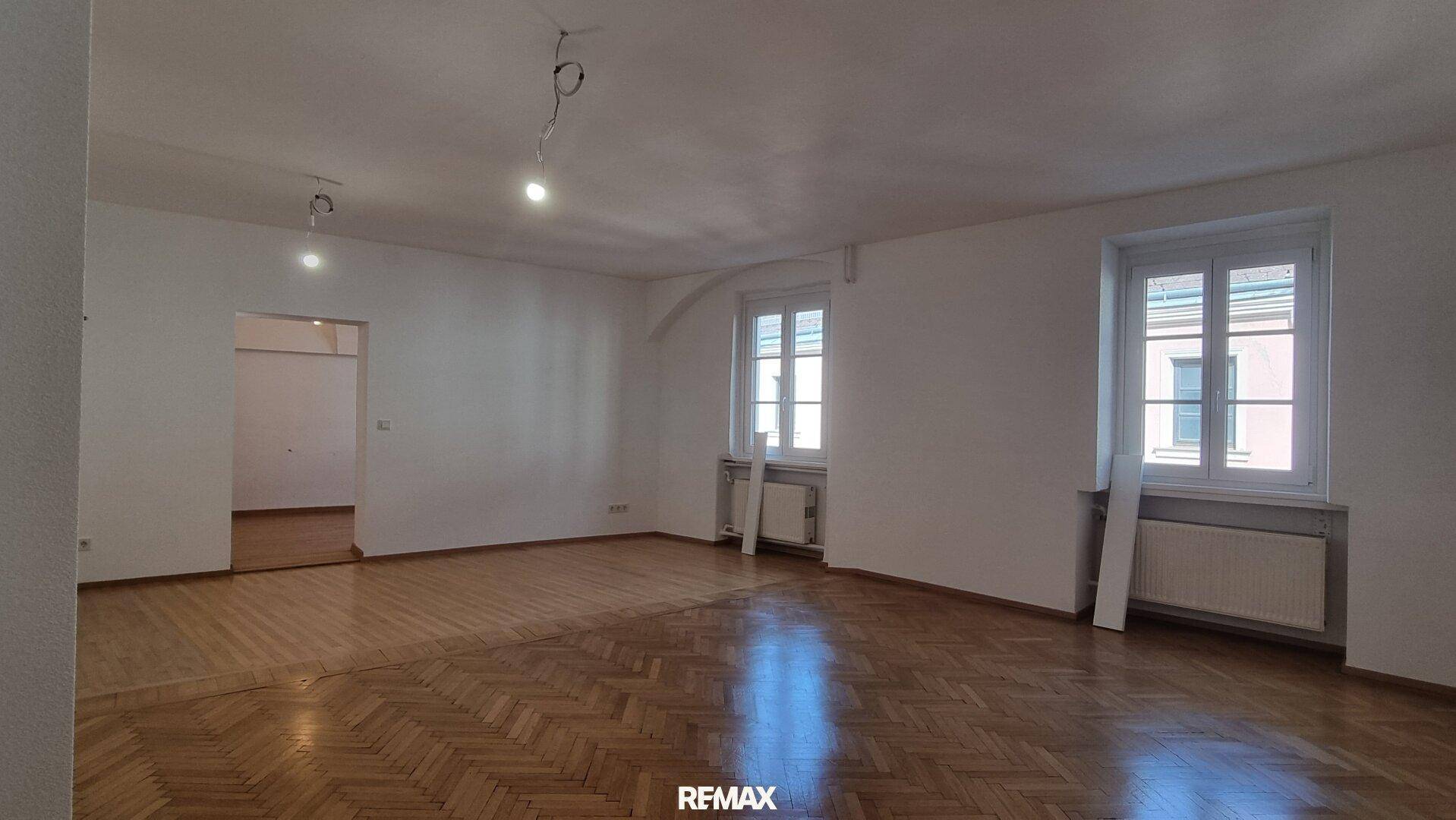 Geräumiges Wohnzimmer mit Fischgrätparkett und großen Fenstern, bietet viel Tageslicht.