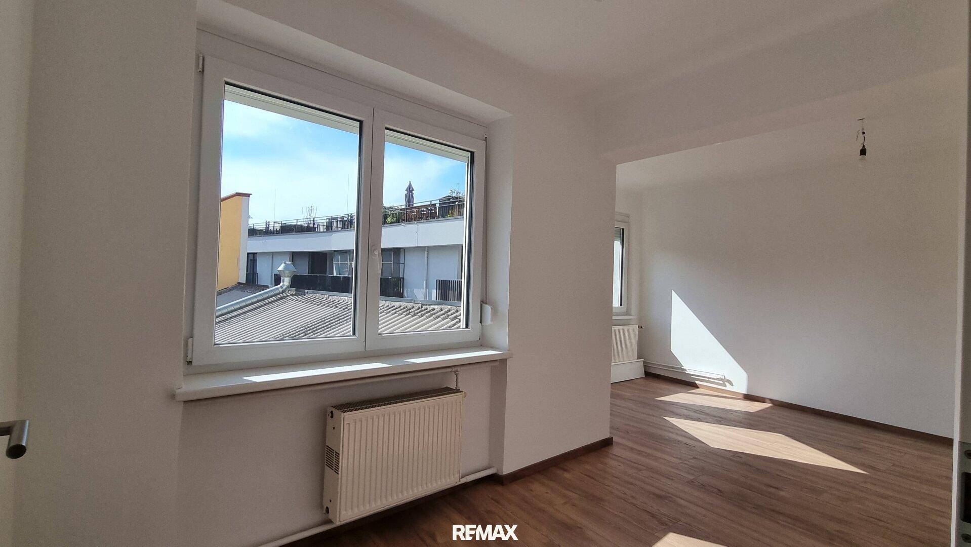 Heller Raum mit großem Fenster und Blick auf umliegende Gebäude und Dächer.