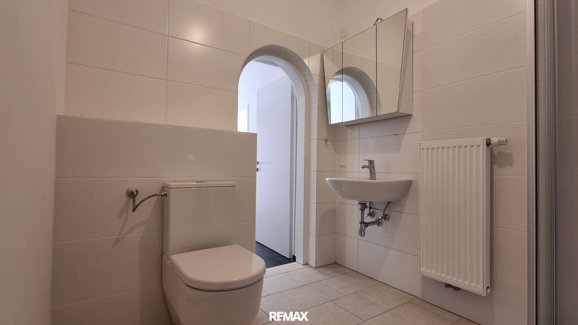 Helles Badezimmer mit WC, Waschbecken und Spiegelschrank, durch einen Bogen verbunden.