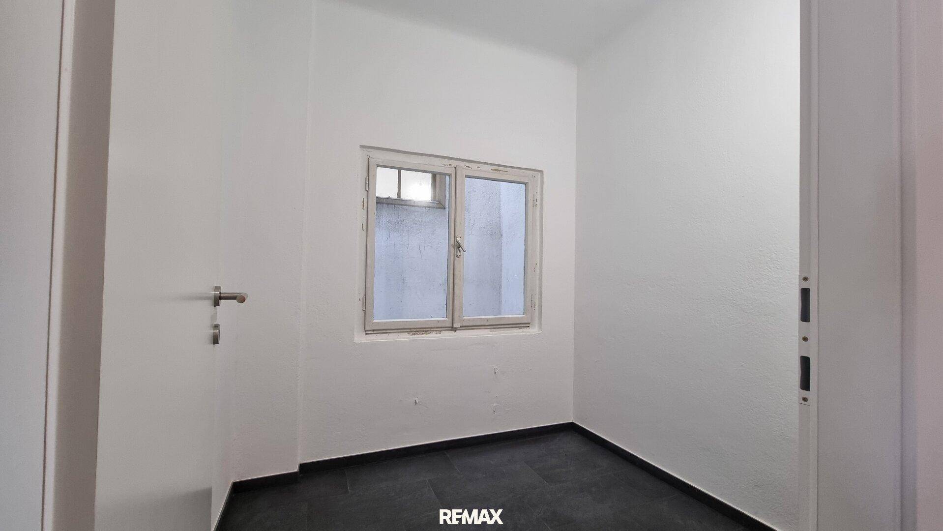Kleiner, heller Raum mit dunklem Fliesenboden und Fenster, ideal als Abstell- oder Arbeitszimmer.