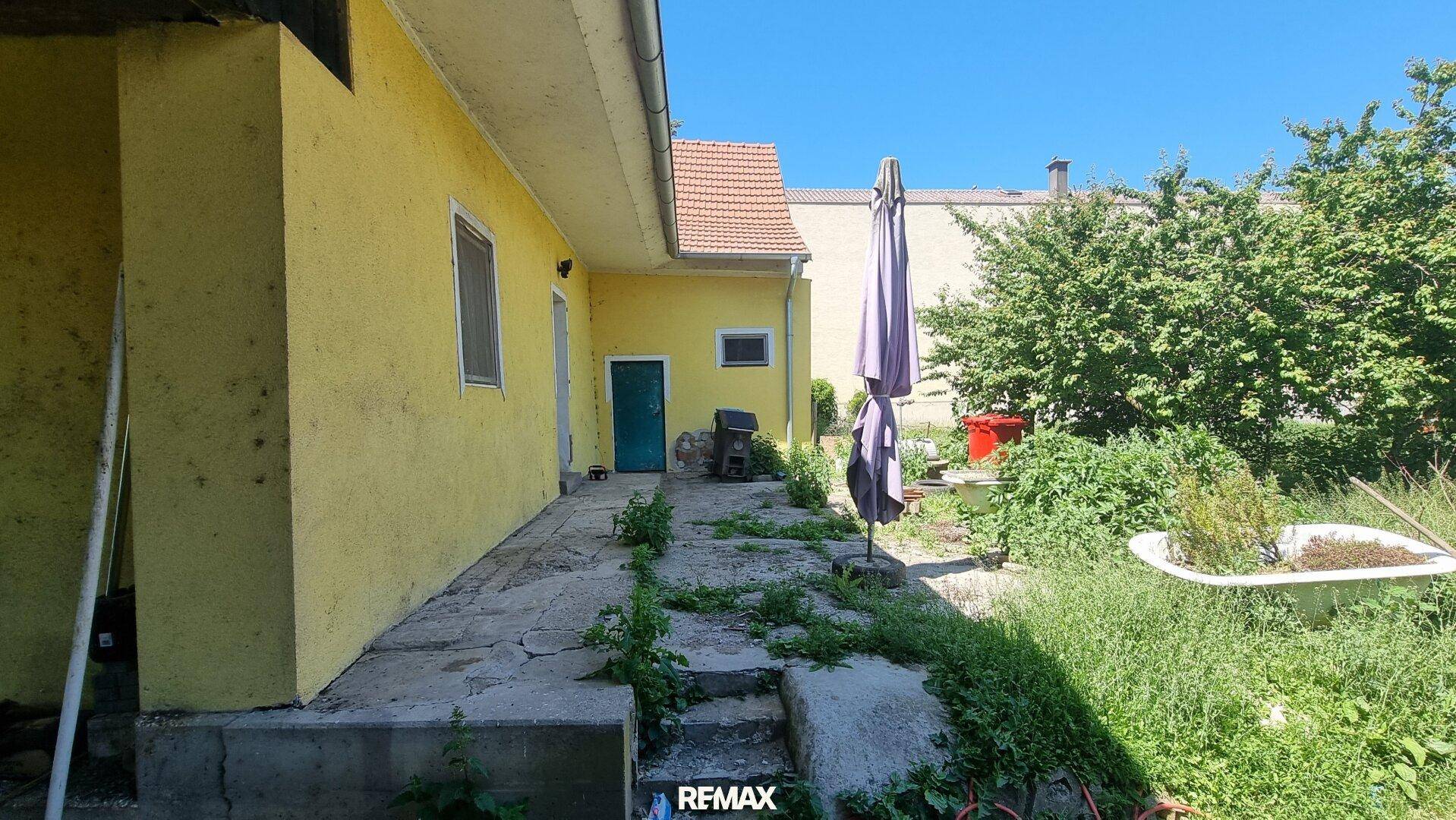 Seite eines gelben Hauses mit verwildertem Gartenbereich, Betonweg und einem Sonnenschirm im Freien.