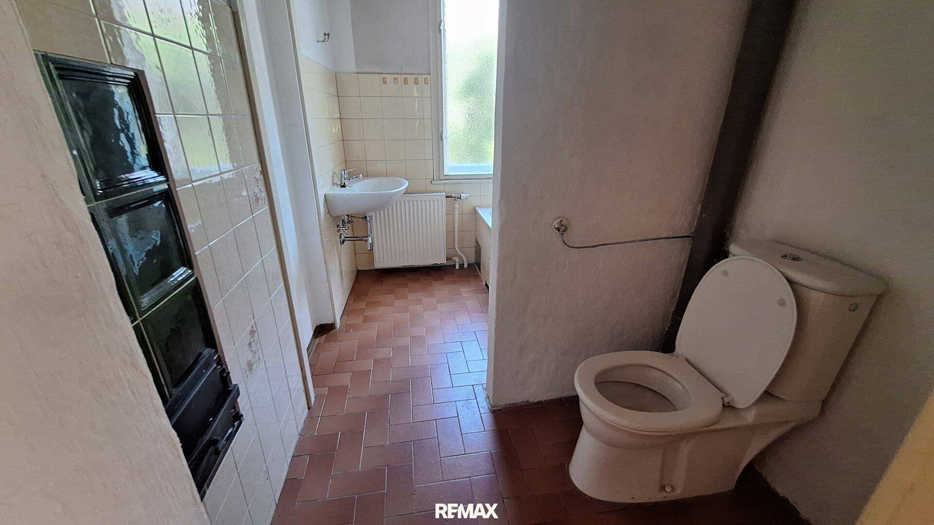 Badezimmer mit Toilette, Waschbecken und Fliesenboden, teilweise mit Kachelofen.