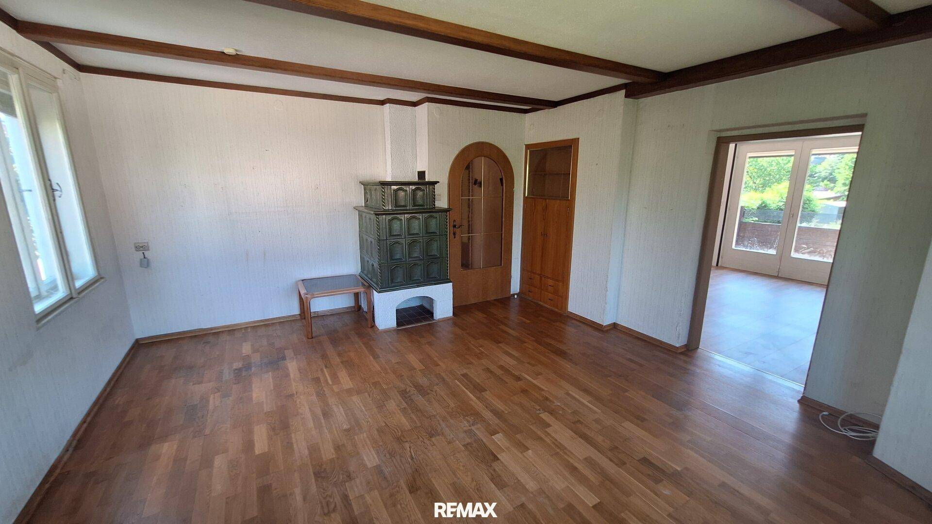 Geräumiges Wohnzimmer mit Holzbalkendecke, Parkettboden und einem traditionellen Kachelofen.