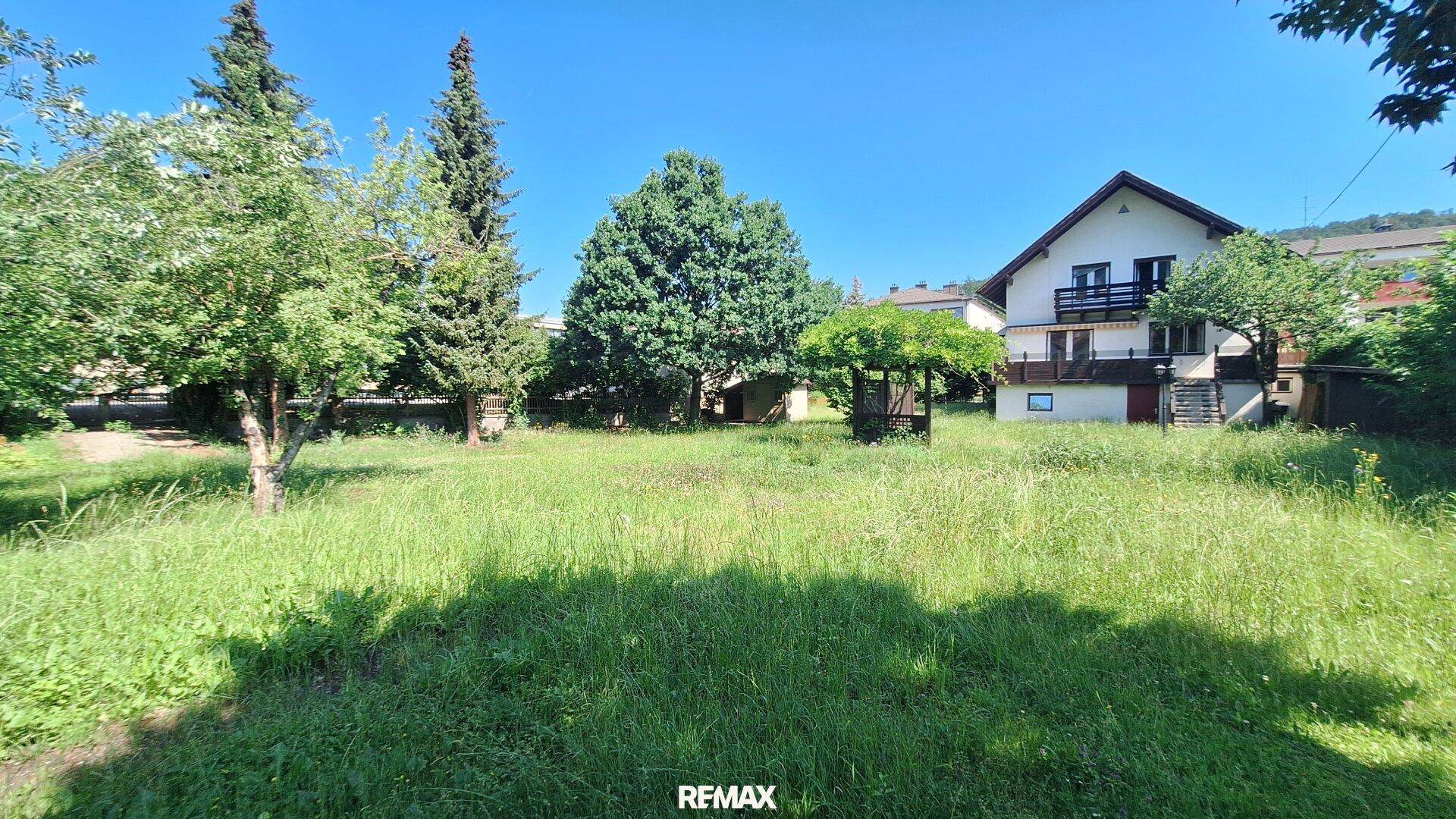 Großer, sonniger Garten mit Rasenfläche, Bäumen und einer kleinen Pergola im Hintergrund.