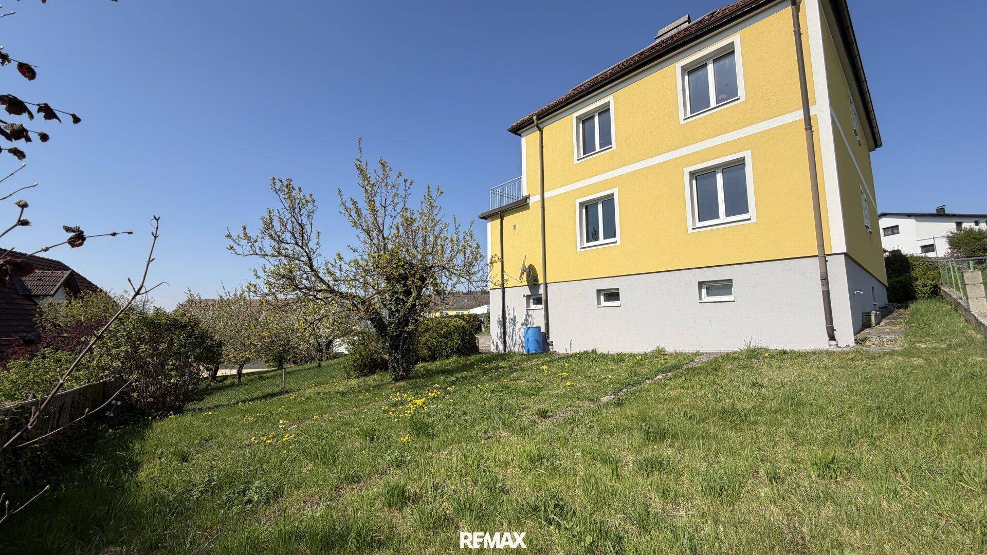 Seitenansicht des gelben Hauses mit Rasenfläche und Bäumen im Sonnenschein.