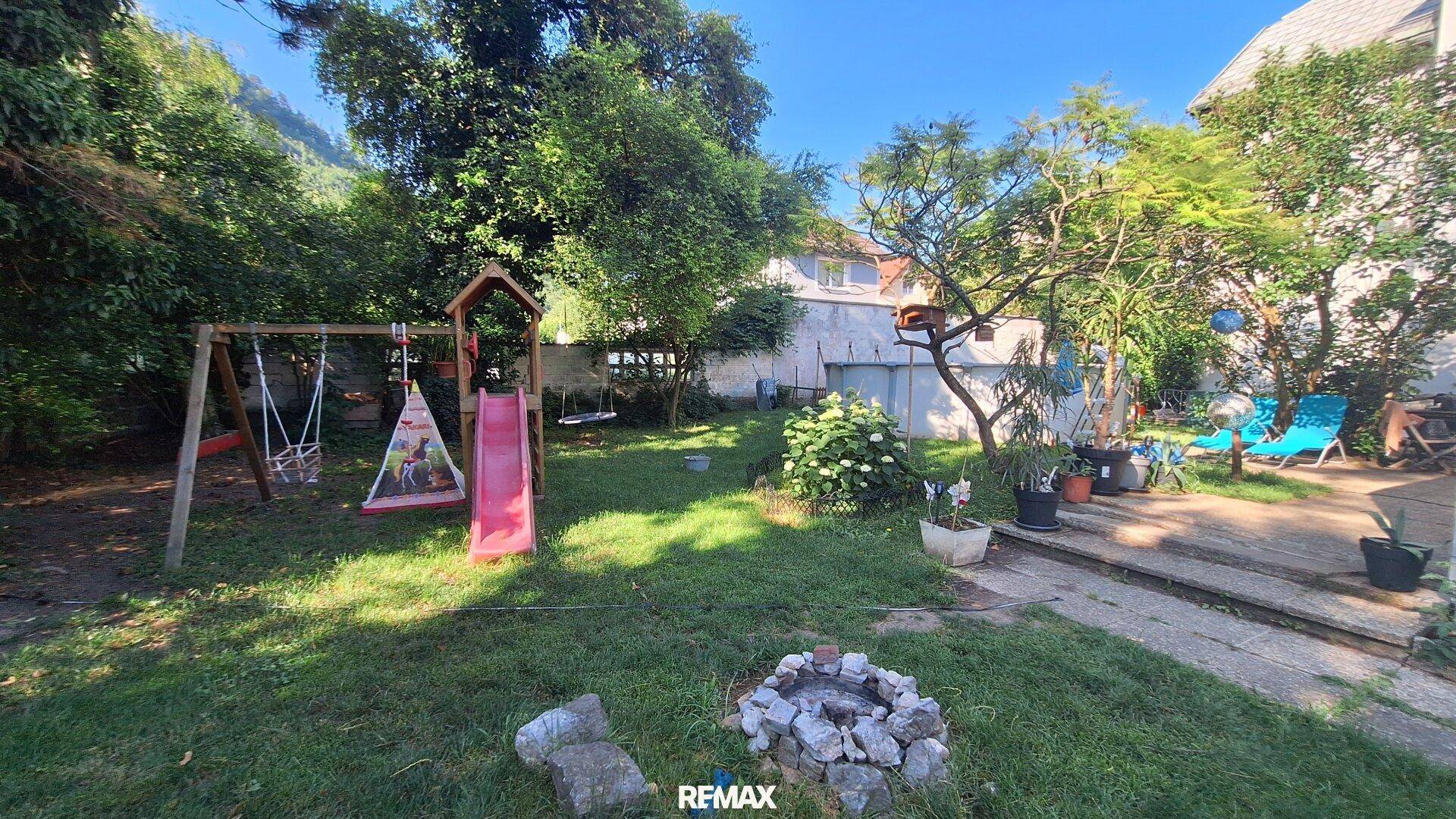 Großer Garten mit Rasenfläche, Spielplatz, Bäumen und einer Feuerstelle.