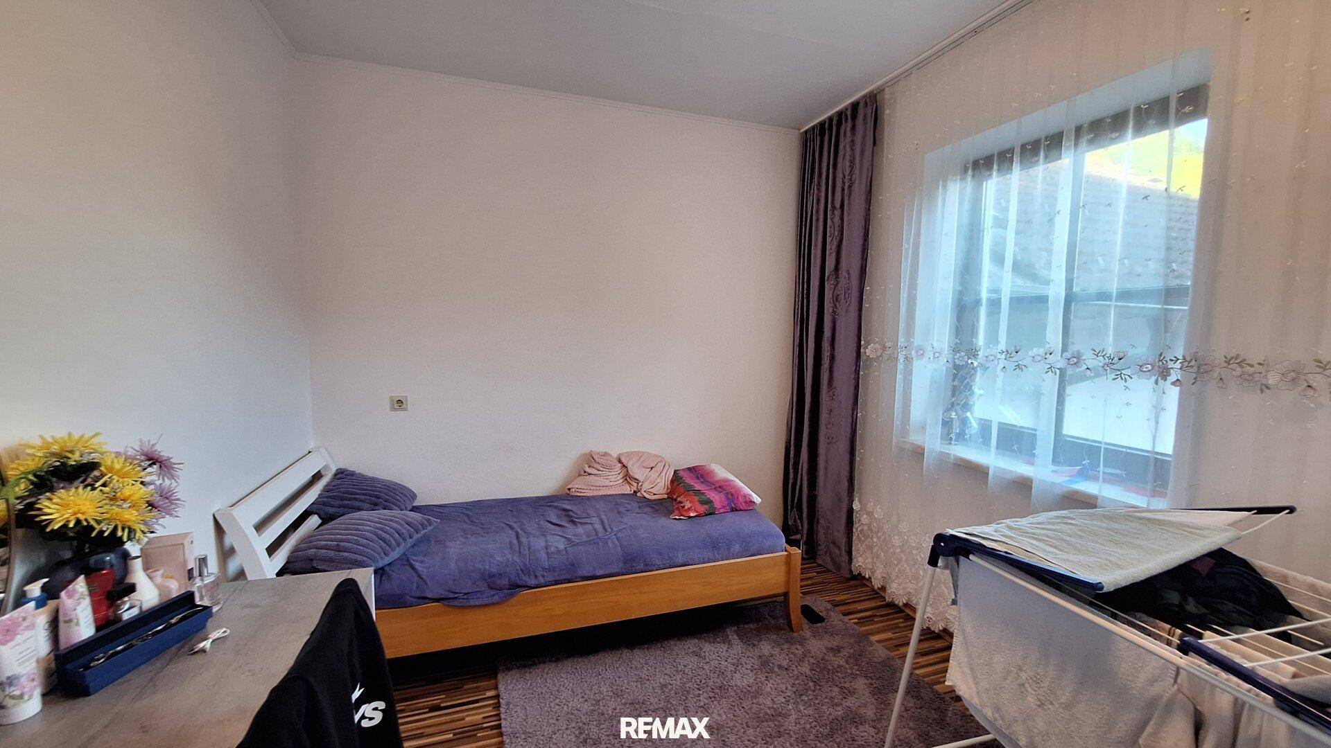 Schlafzimmer mit Einzelbett, Teppich und Fenster mit lila Vorhängen.