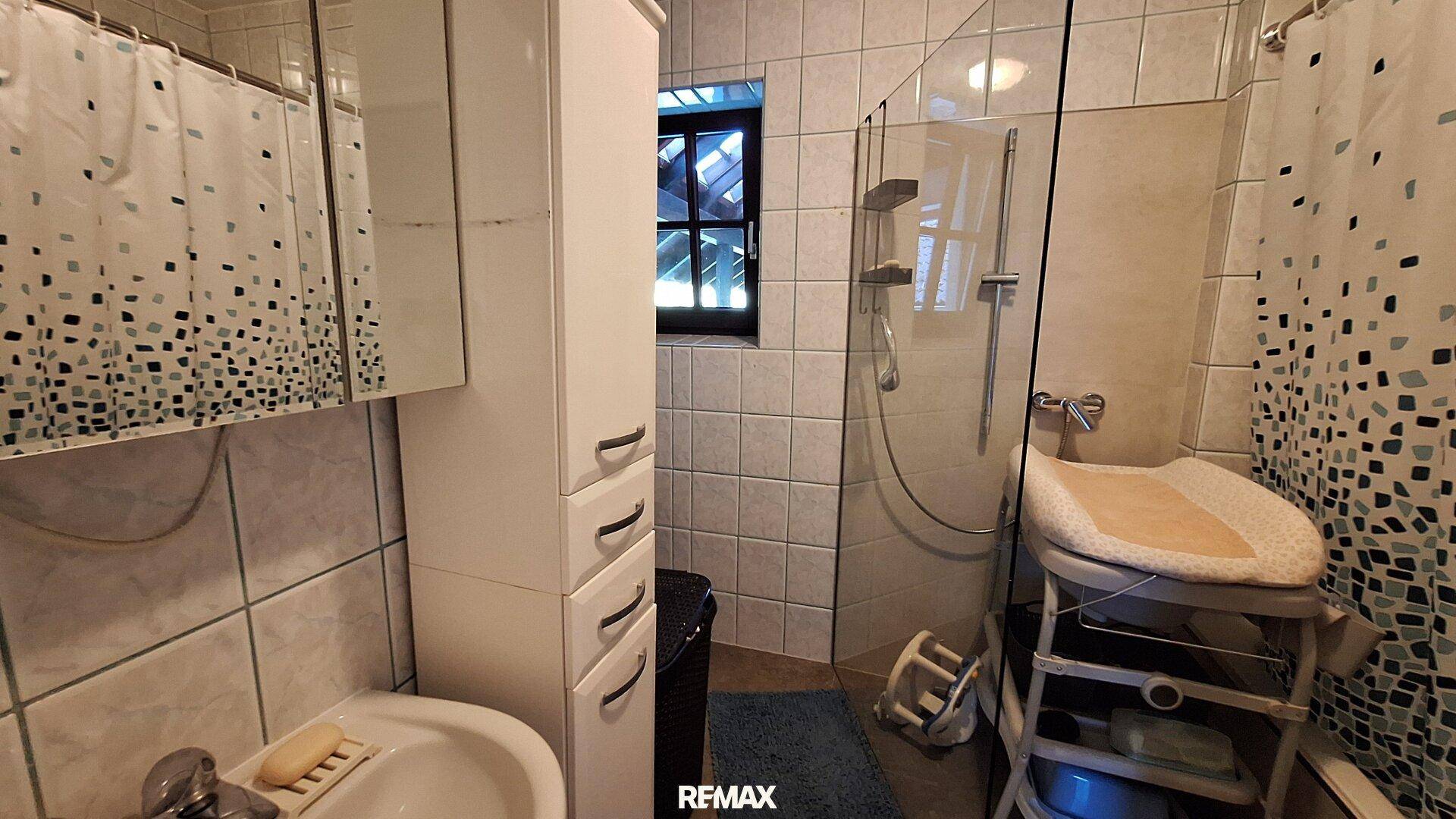 Gefliestes Badezimmer mit Dusche, Waschbecken und Fenster im Dachschrägenbereich.