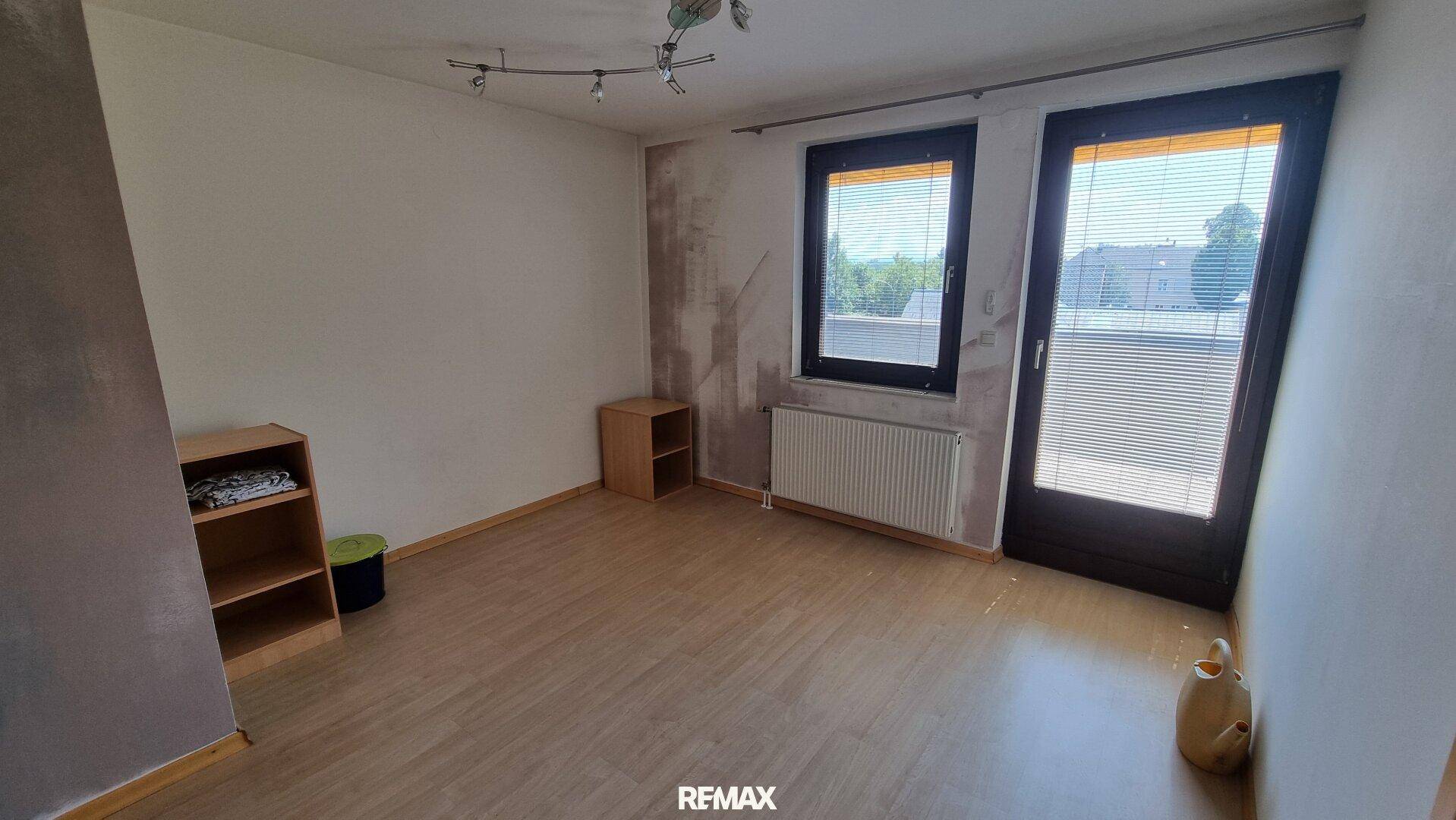 Leerer Raum mit Laminatboden, Fenster und Zugang zu einem weiteren Bereich, renovierungsbedürftig.
