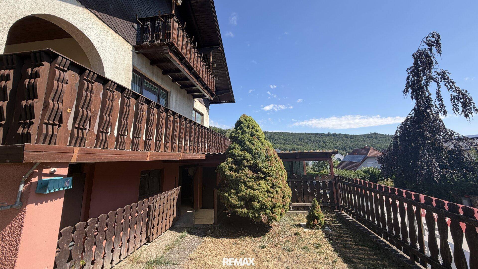 Seitenansicht des Hauses mit traditionellem Holzbalkon und einem kleinen, ungepflegten Gartenbereich.