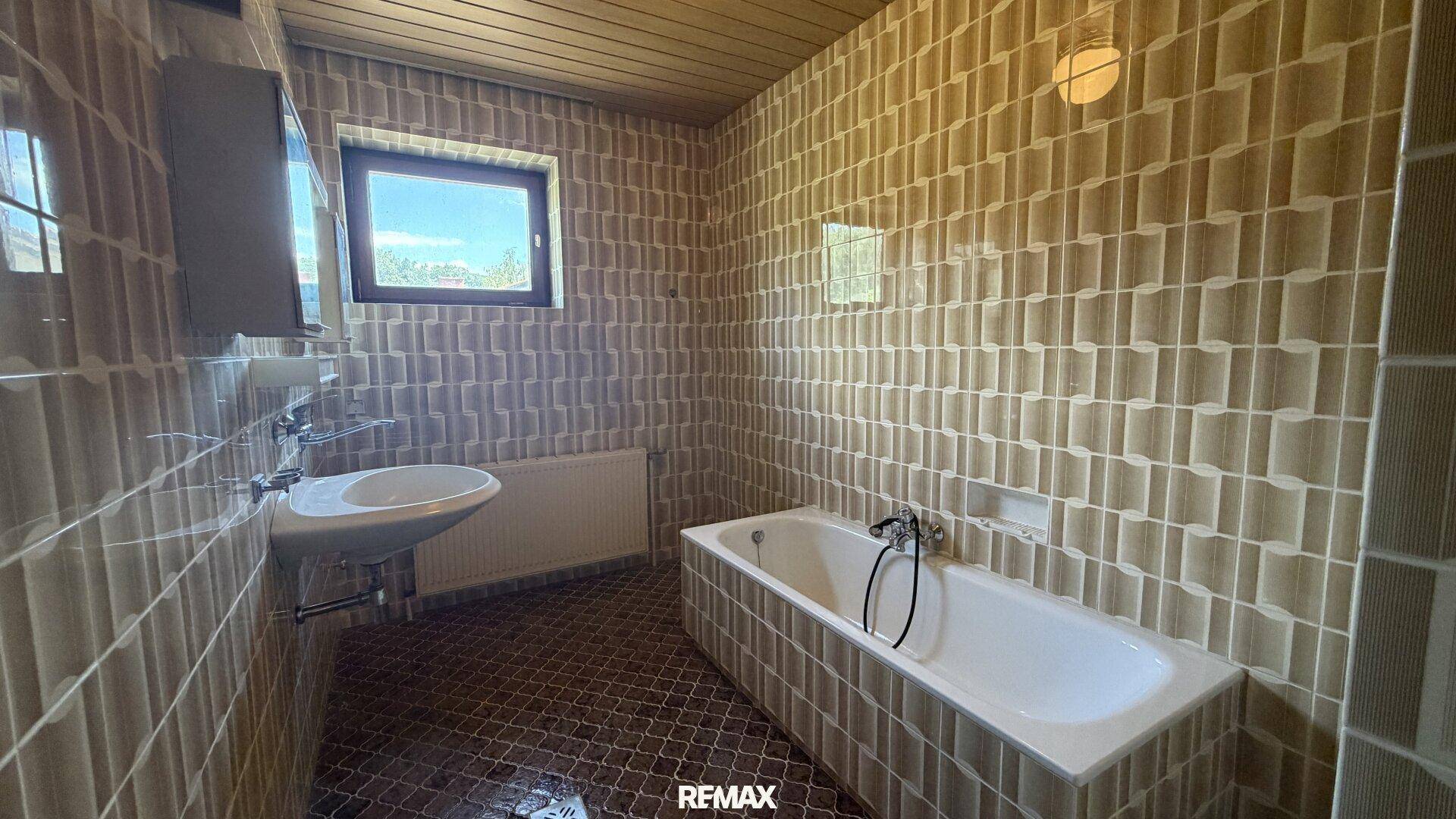 Badezimmer mit beigefarbenen Fliesen, Badewanne, Waschbecken und einem Fenster für Tageslicht.