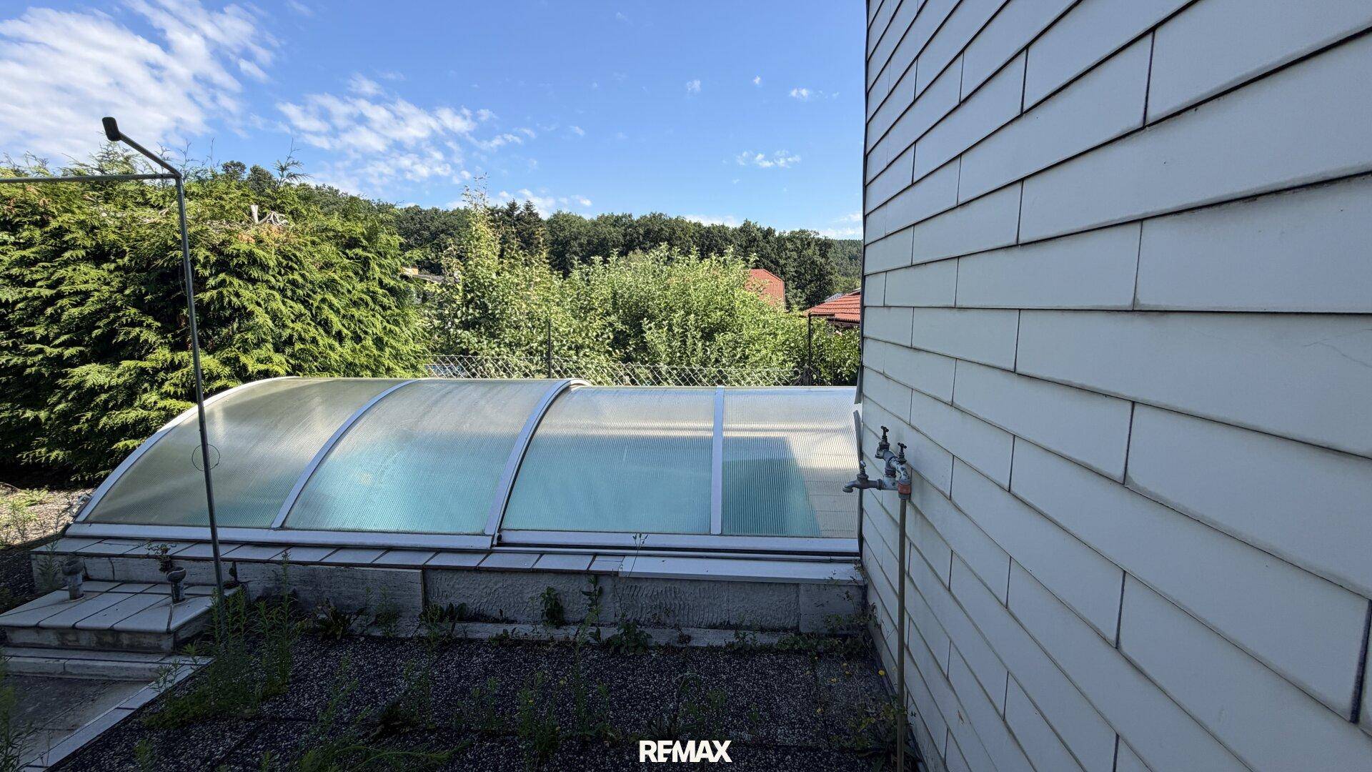 Außenansicht des Hauses mit einem überdachten Pool und Blick ins Grüne unter blauem Himmel.