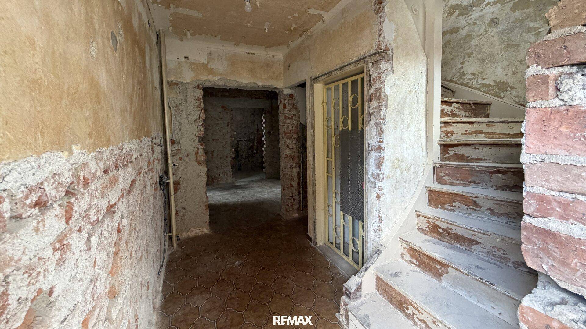 Flur im Rohbauzustand mit freigelegten Ziegelwänden und einer alten Holztreppe.