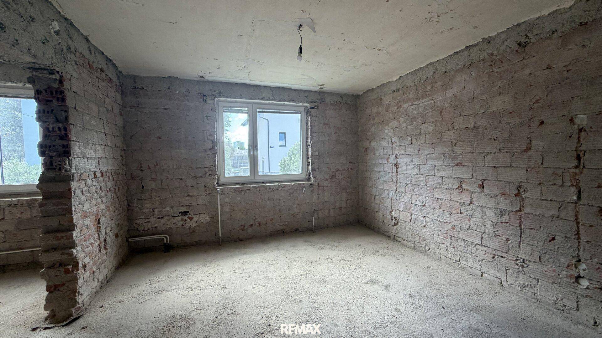 Raum im Rohbau mit freigelegten Ziegelwänden und einem Fenster, bereit für die Renovierung.