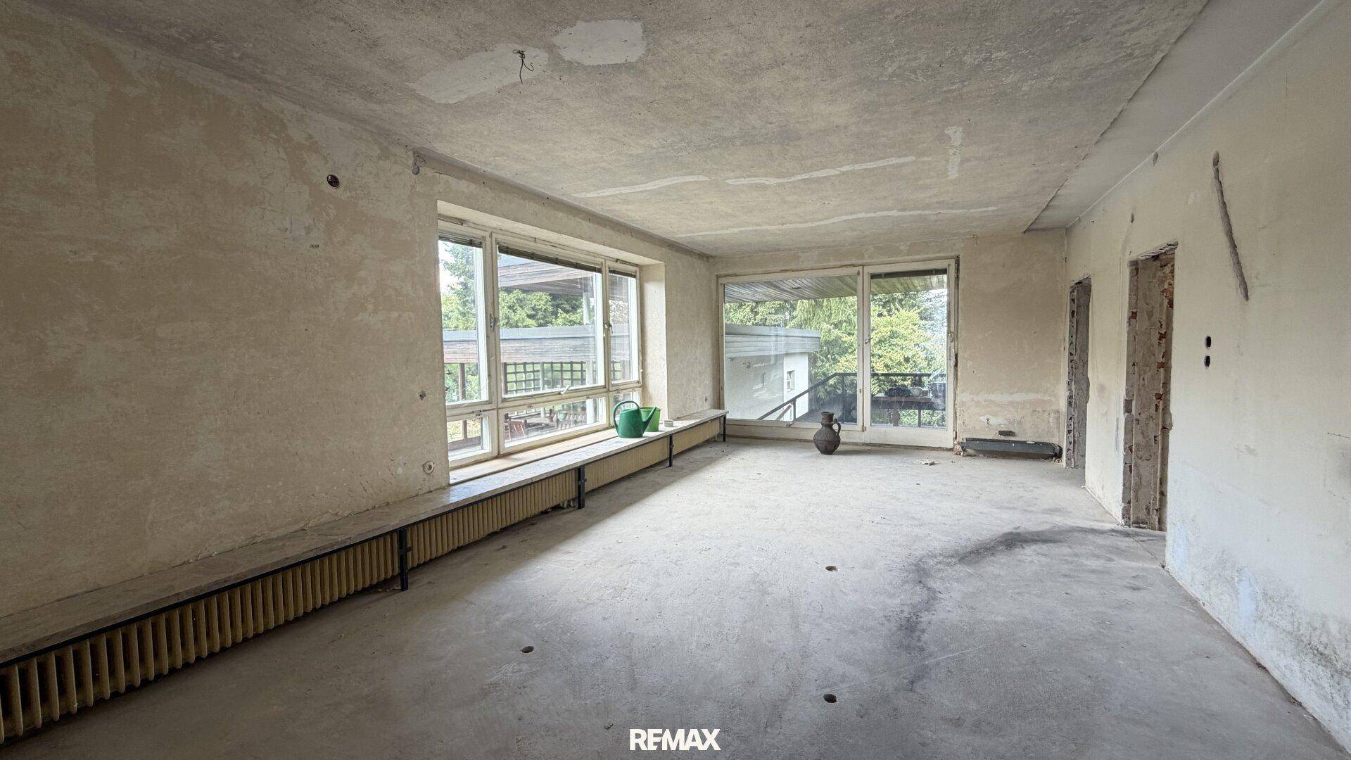 Großer Raum im Rohbau mit breiten Fenstern und Blick ins Grüne, ideal für einen Wohnbereich.