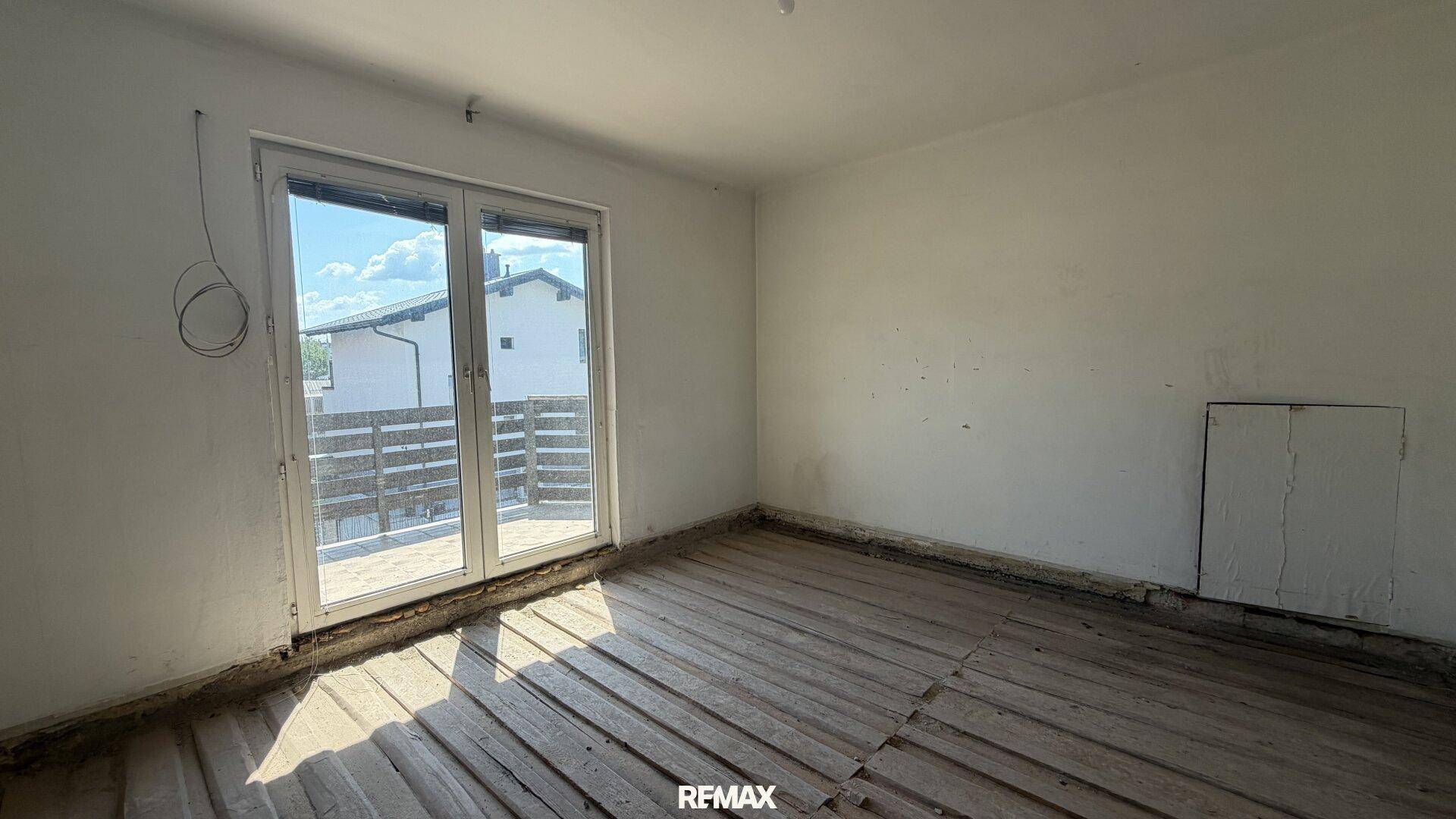 Schlafzimmer im Rohbau mit Zugang zu einem Balkon und sichtbaren Holzdielen auf dem Boden.