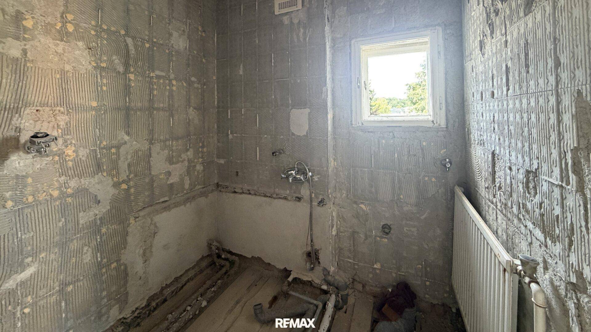 Badezimmer im Rohbau mit freigelegten Wänden, Fenster und sichtbaren Installationen.