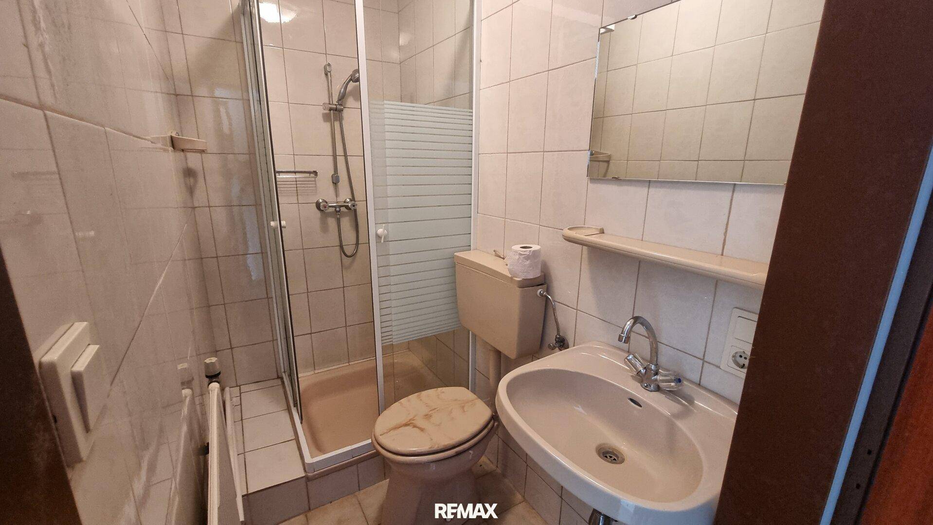 Badezimmer mit beigefarbenen Fliesen, Dusche, WC und Waschbecken in älterem Zustand.