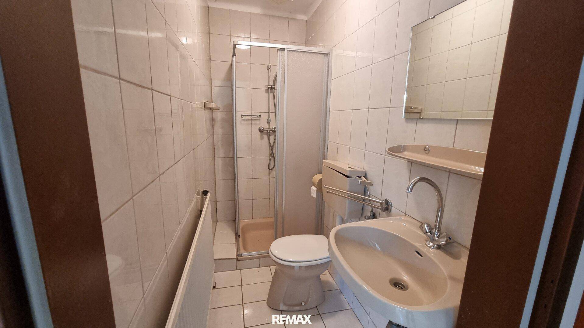 Badezimmer mit beigefarbenen Fliesen, Dusche, WC und Waschbecken in älterem Zustand.