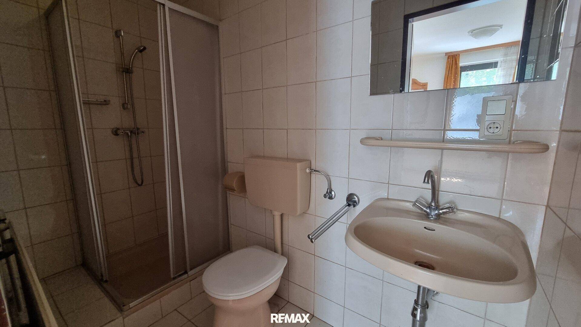 Badezimmer mit beigefarbenen Fliesen, Dusche, WC und Waschbecken in älterem Zustand.