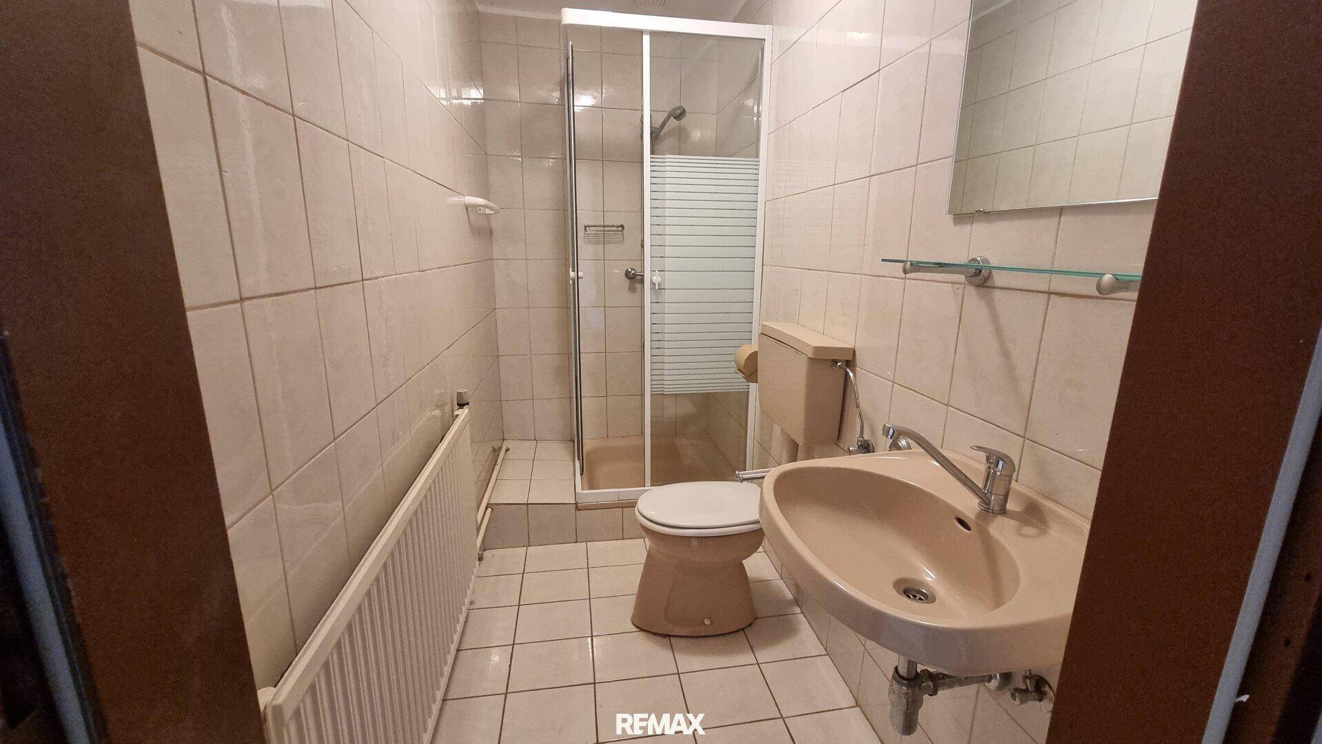 Badezimmer mit beigefarbenen Fliesen, Dusche, WC und Waschbecken in älterem Zustand.