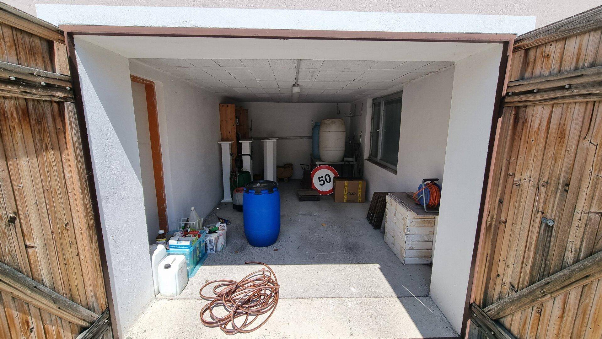 Geräumige Garage mit offenem Holztor, Betonboden und Stauraum für verschiedene Gegenstände.