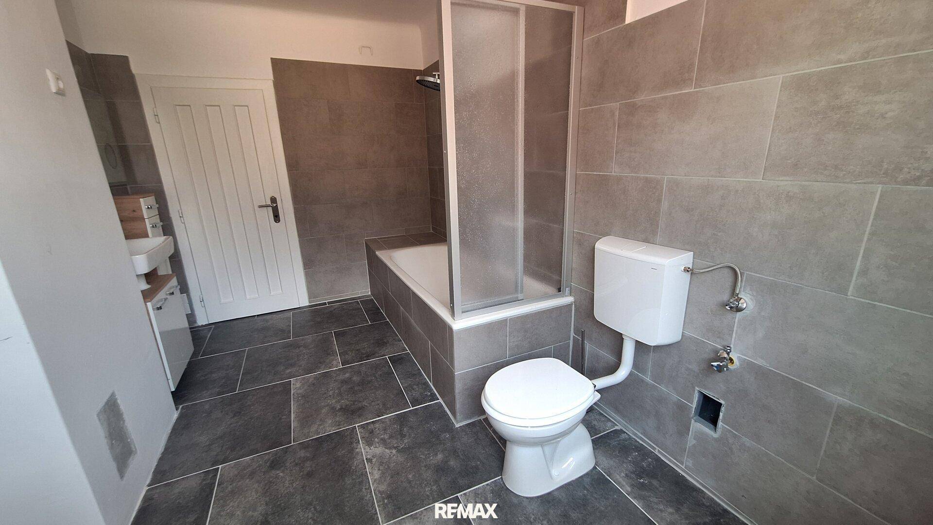 Funktionales Badezimmer mit grauen Fliesen, einer Badewanne mit Duschabtrennung und einer Toilette.