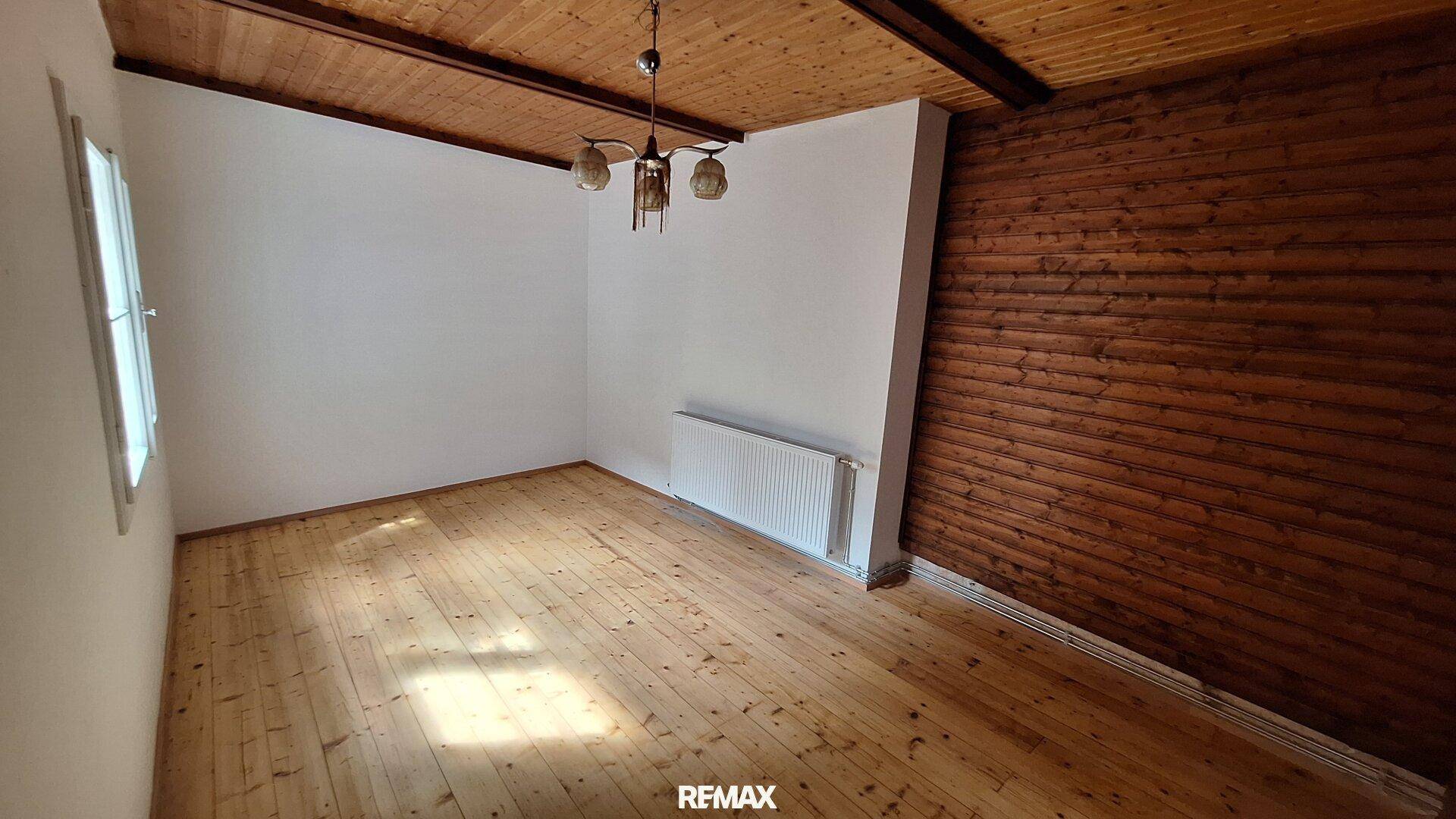 Geräumiges Zimmer mit Holzboden, einer Holzwandvertäfelung und sichtbaren Deckenbalken, unmöbliert.