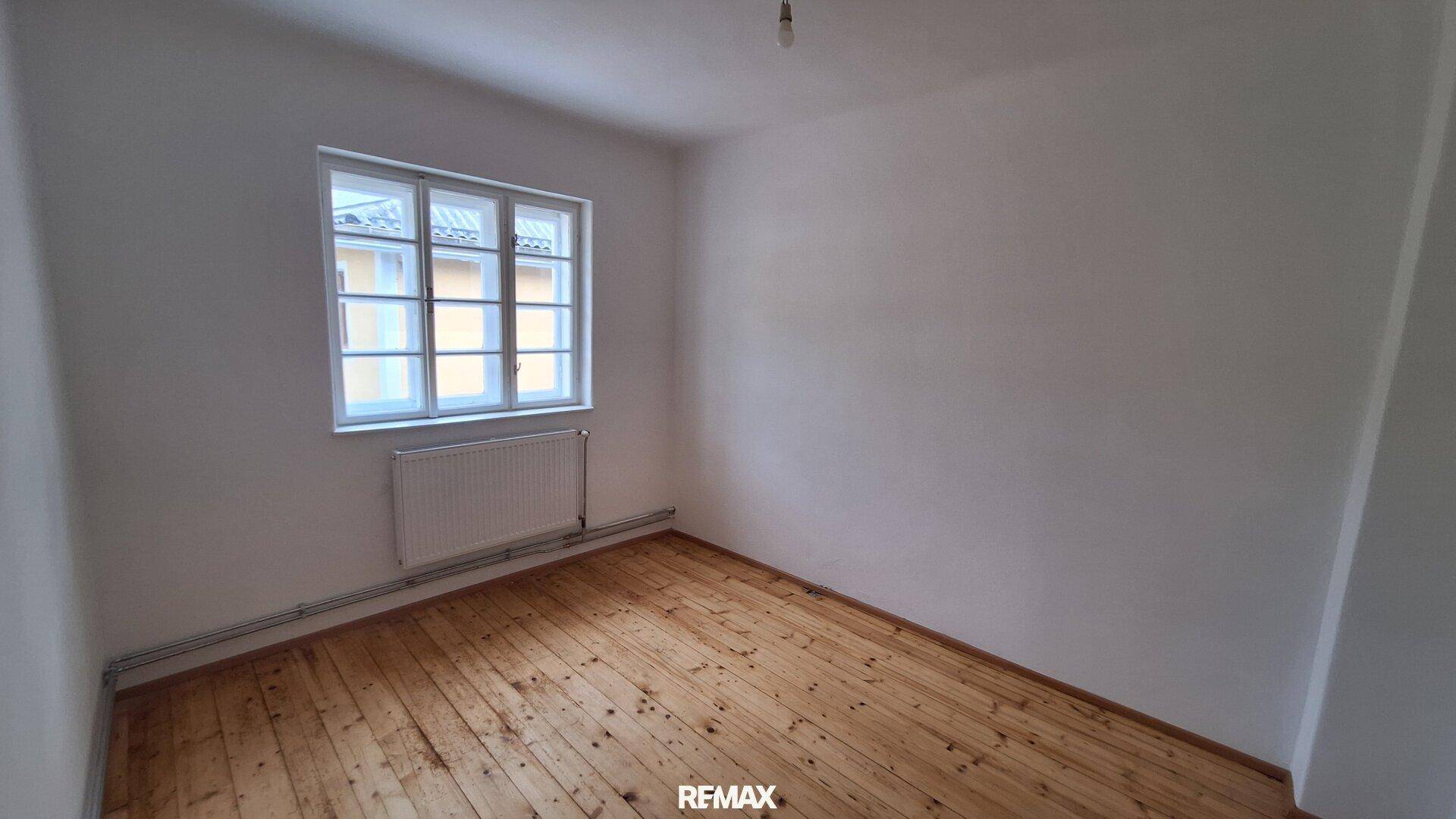 Helles, unmöbliertes Zimmer mit Holzboden, einem großen Fenster und einem Heizkörper an der Wand.