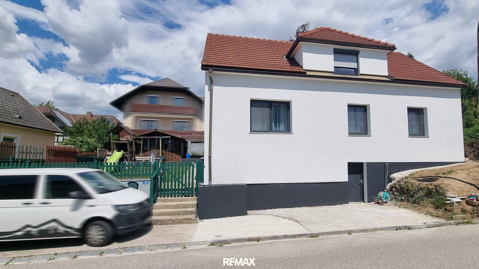 Seitenansicht des Hauses von der Straße mit weißer Fassade, rotem Ziegeldach und dunklem Sockel.