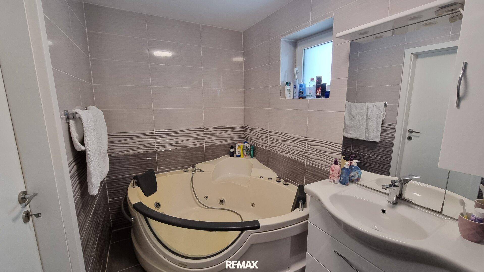 Modernes Badezimmer mit Eck-Whirlpool, hellen Fliesen und einem kleinen Fenster für Tageslicht.