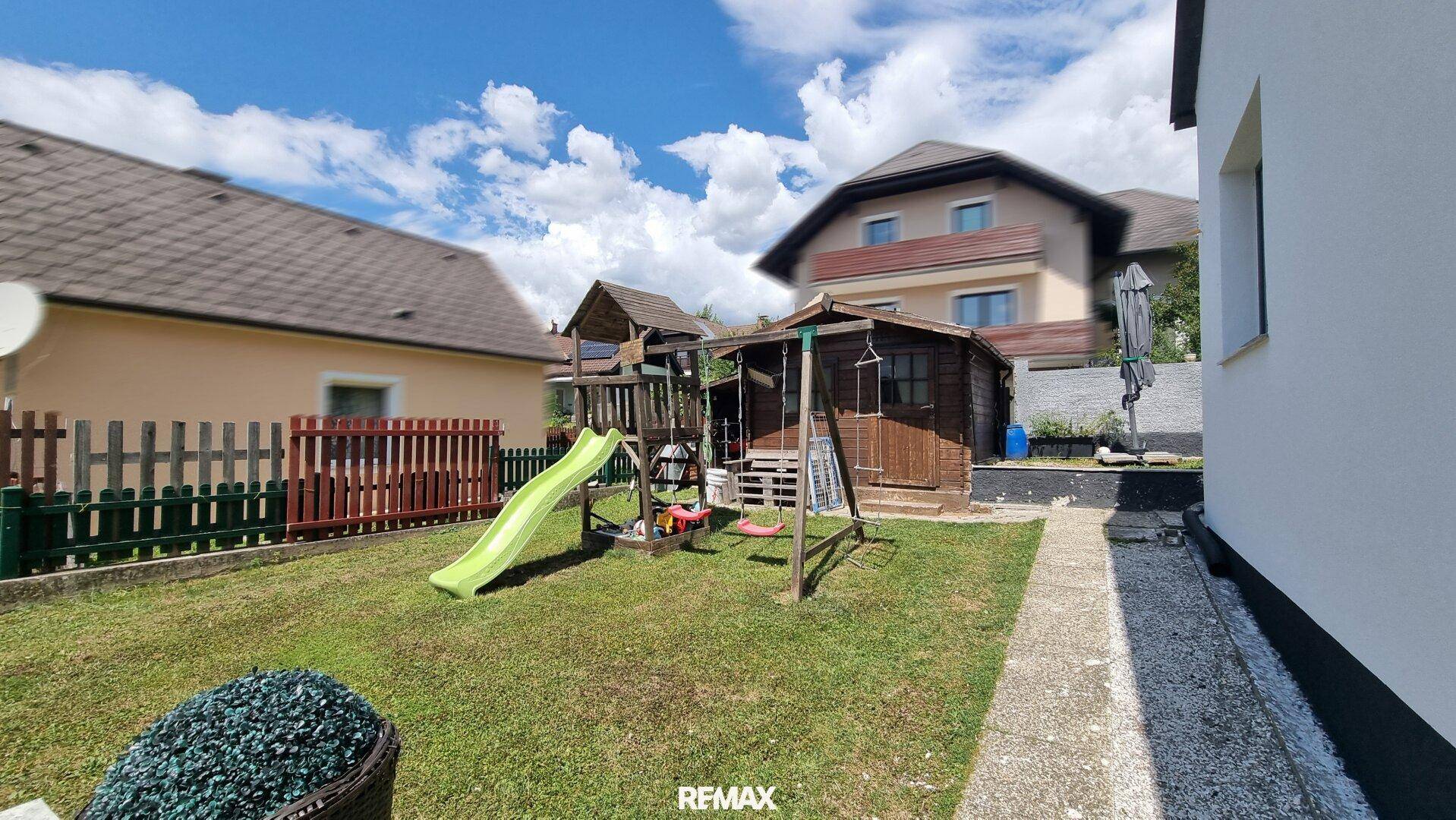 Grüner Gartenbereich mit Kinderspielplatz, Rutsche, Schaukeln und einem kleinen Holzschuppen.