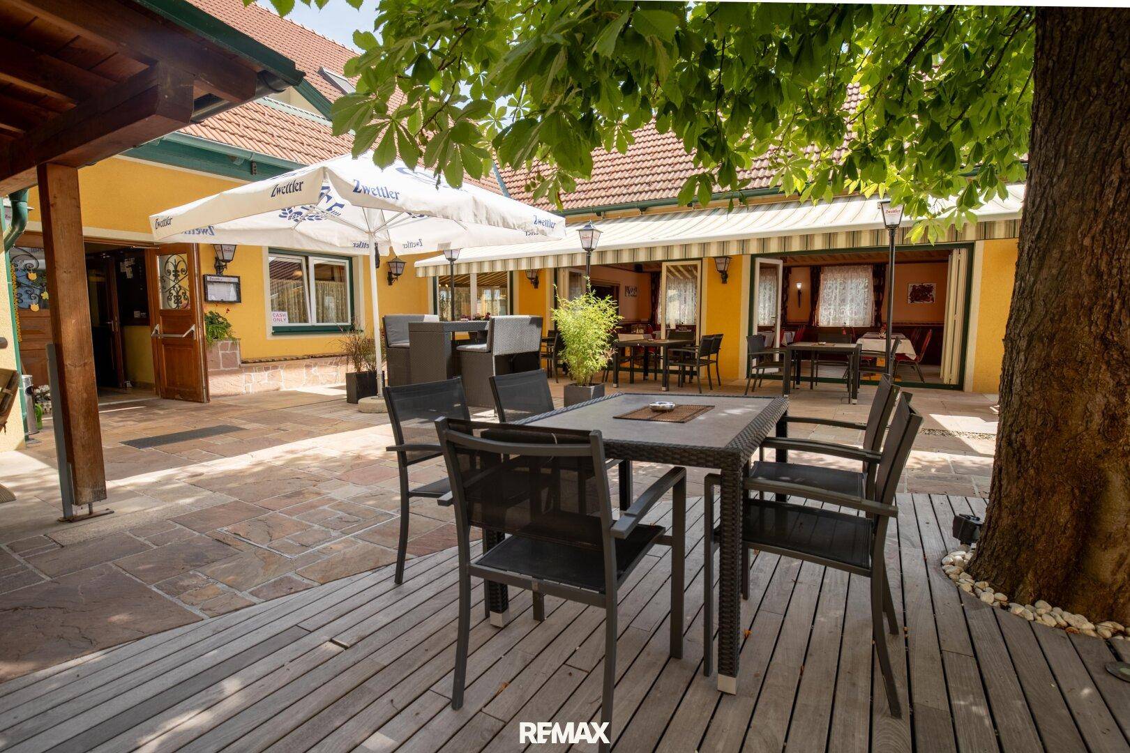 Schattige Terrasse mit Sitzgelegenheiten unter einem großen Baum und Sonnenschirm, perfekt für warme Tage.