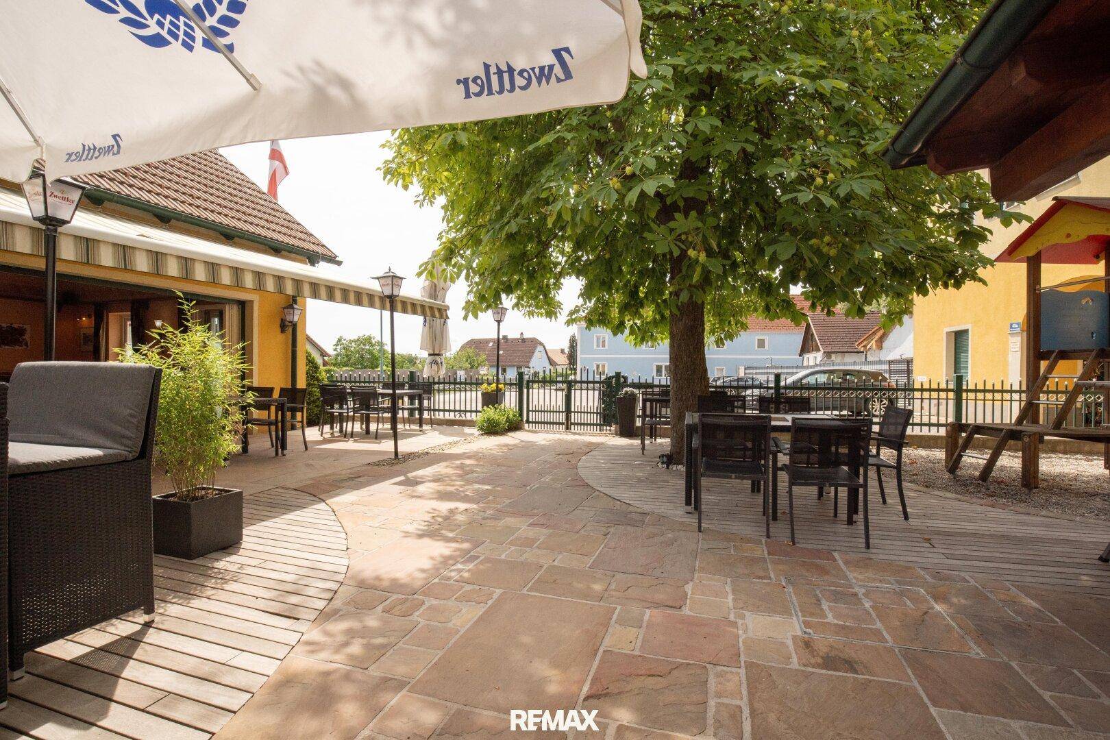 Große gepflasterte Terrasse mit Sitzgelegenheiten und einem Spielbereich für Kinder unter schattigen Bäumen.