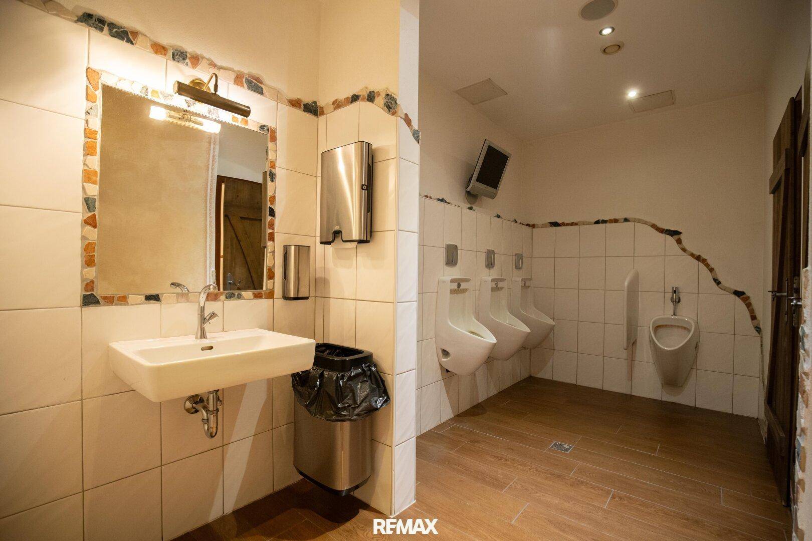 Herren-WC mit mehreren Urinalen, Waschbecken und gefliesten Wänden in hellem Design.