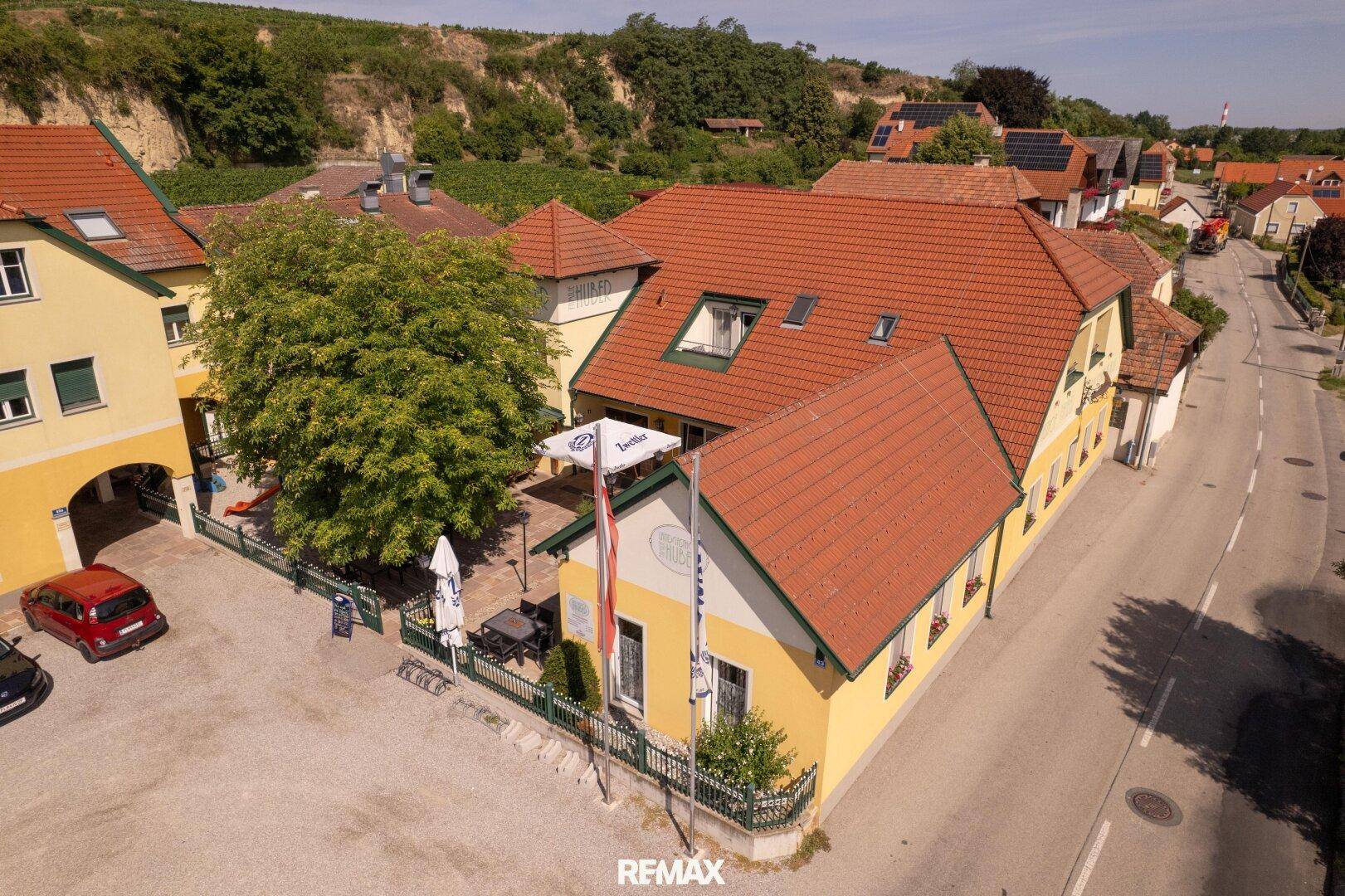 Luftaufnahme eines Gasthofs mit rotem Ziegeldach, gelber Fassade und angrenzendem Weinberg in ländlicher Umgebung.