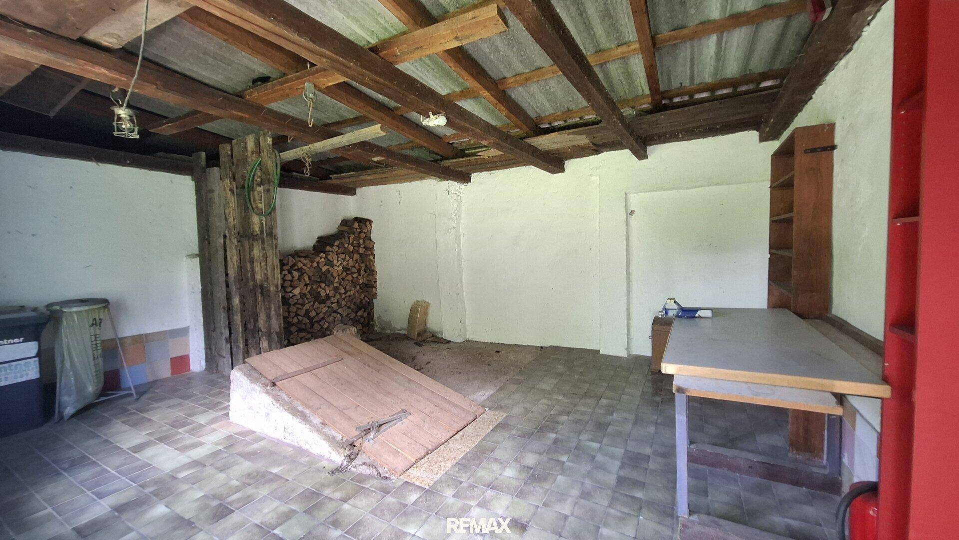 Kellerraum mit sichtbaren Holzbalken, Fliesenboden und gelagertem Brennholz.