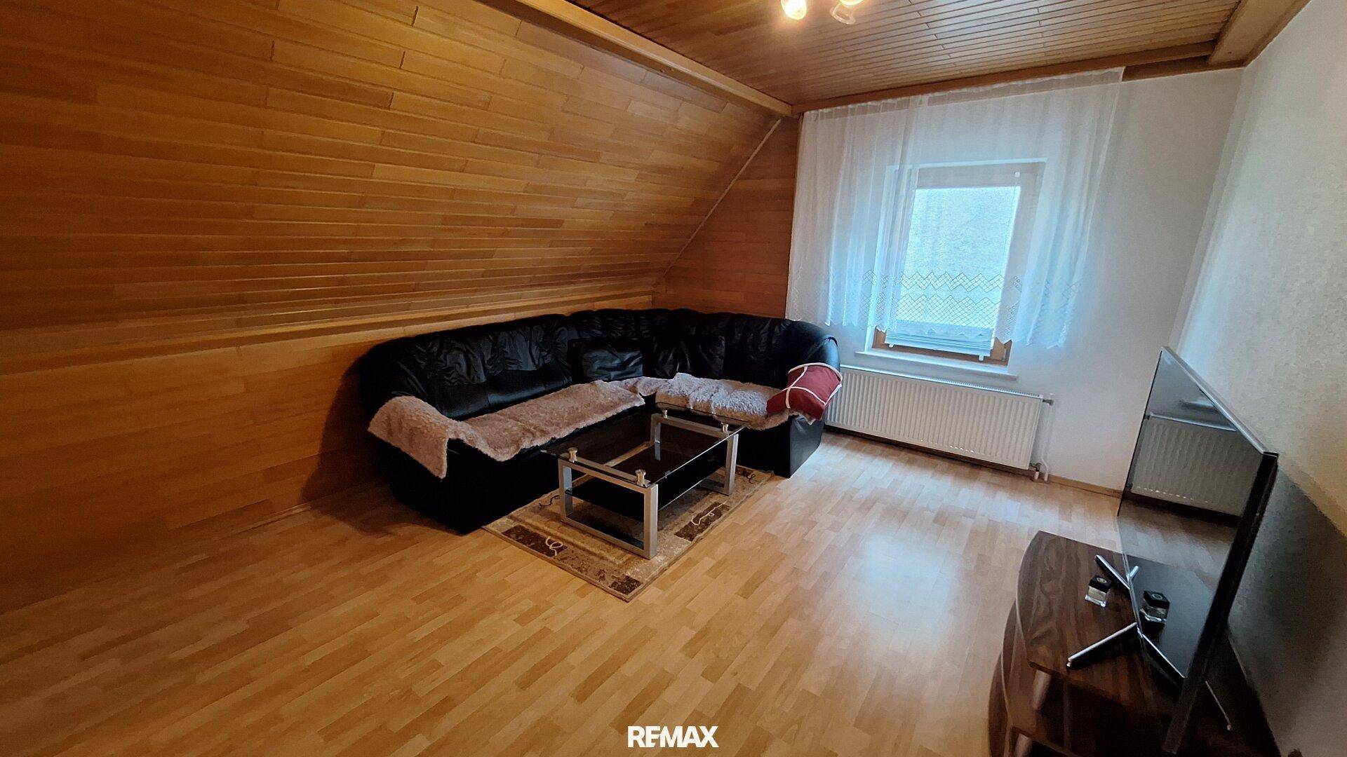 Wohnzimmer im Dachgeschoss mit Holzbalkendecke, Parkettboden und einem großen schwarzen Sofa.
