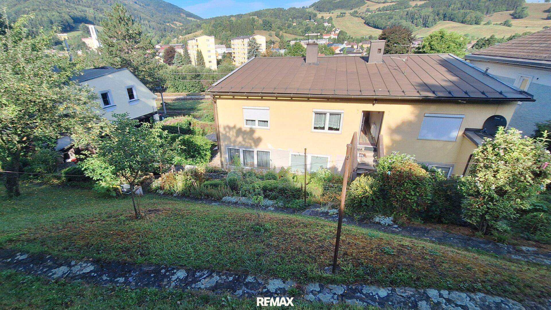 Haus mit Garten auf einem Hügel, umgeben von grüner Landschaft und weiteren Gebäuden.