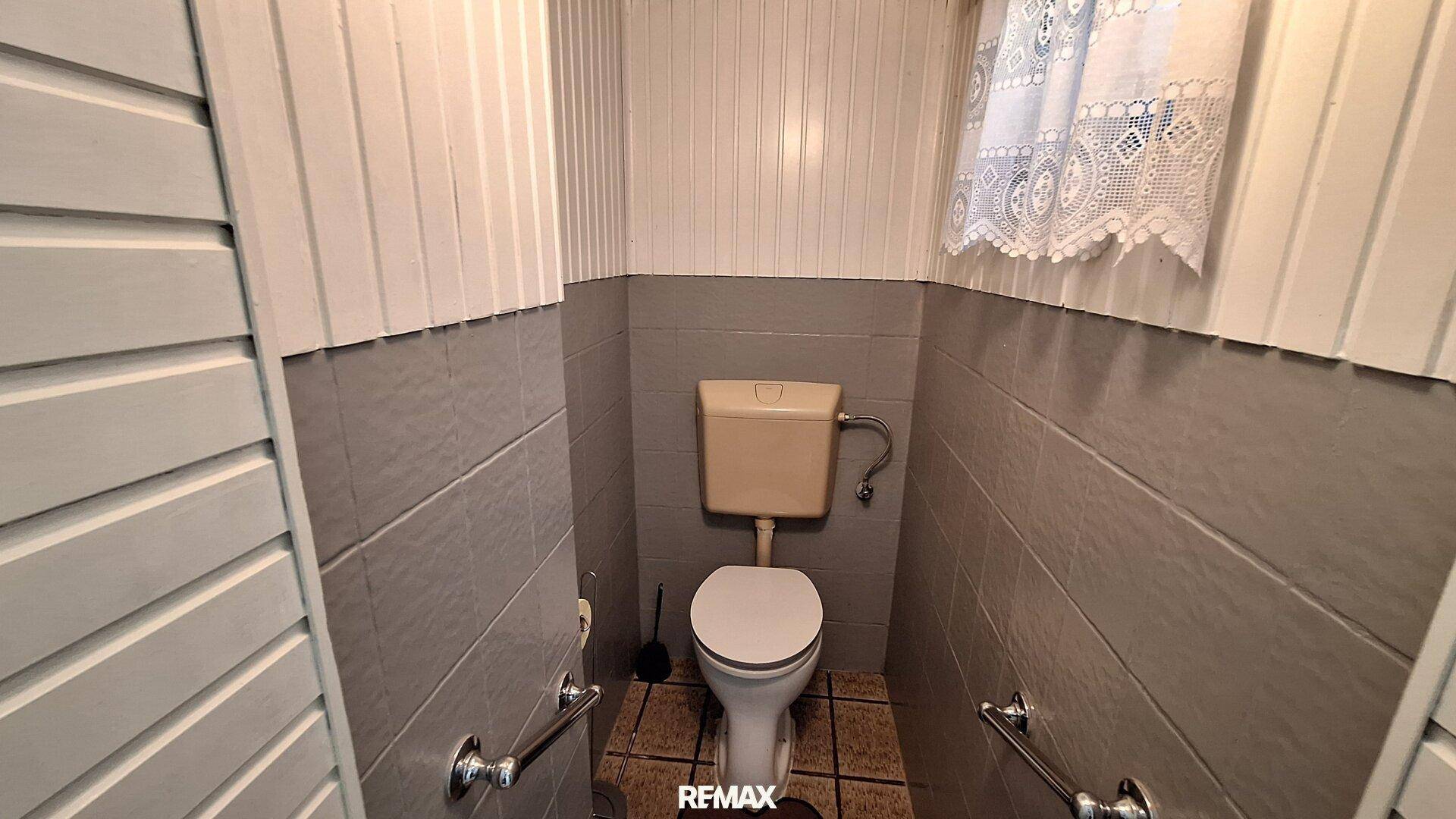 Kleines Gäste-WC mit grauen Fliesen, Toilette und Haltegriffen.