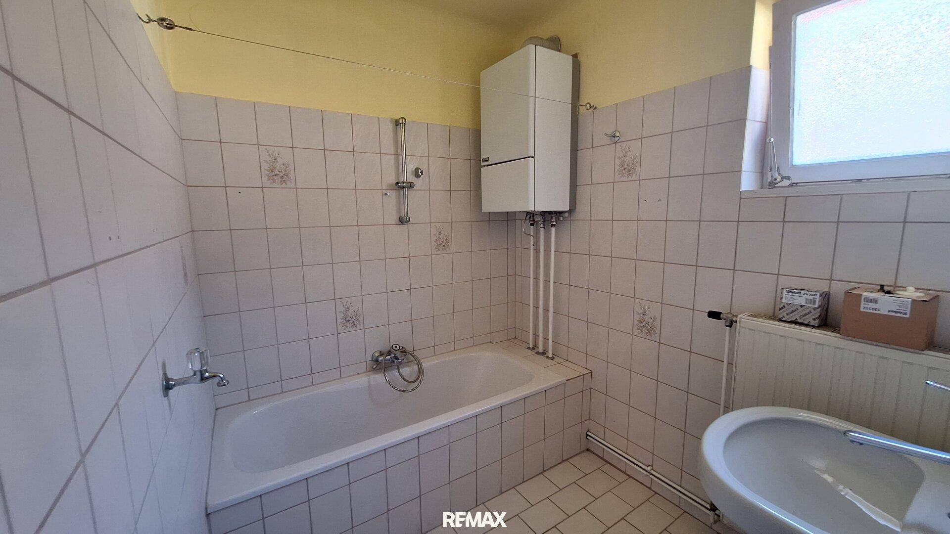 Badezimmer mit weißen Fliesen, Badewanne, Boiler und Fenster.