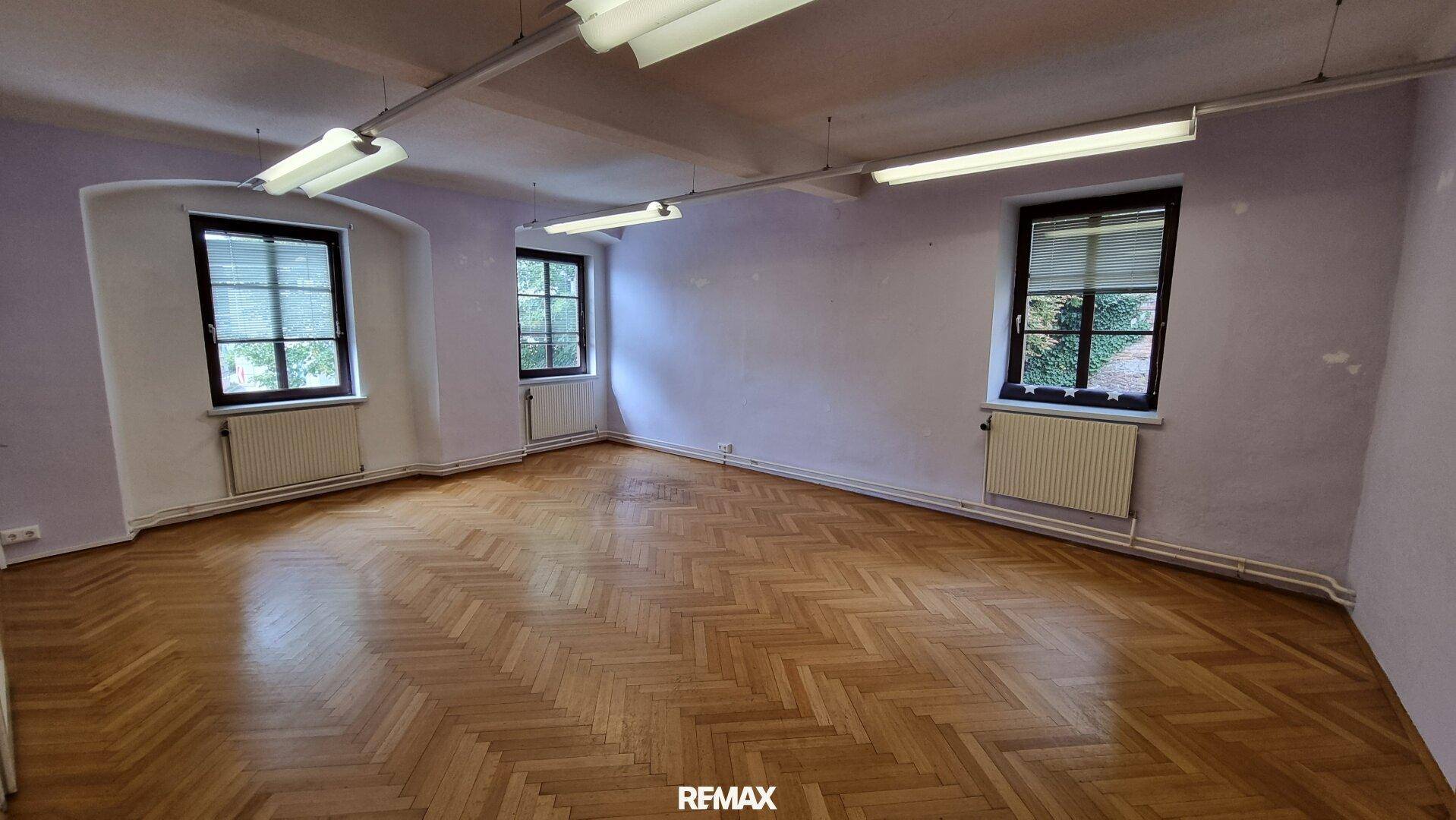 Großer Raum mit Fischgrätparkett und drei Fenstern, ideal für Büro oder Wohnzwecke.