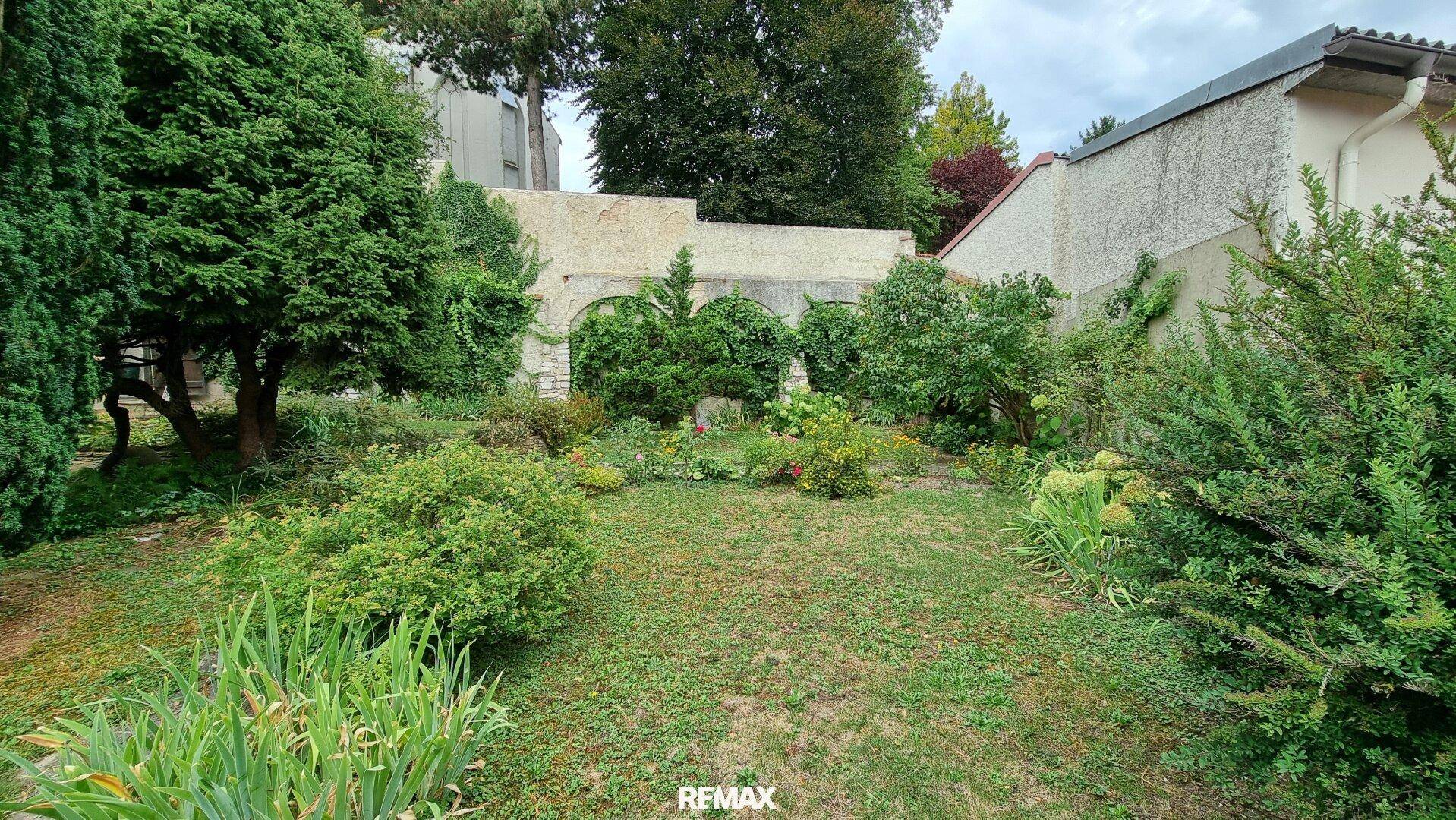 Weitläufiger Garten mit Rasenfläche und verschiedenen Sträuchern, umgeben von einer Mauer.