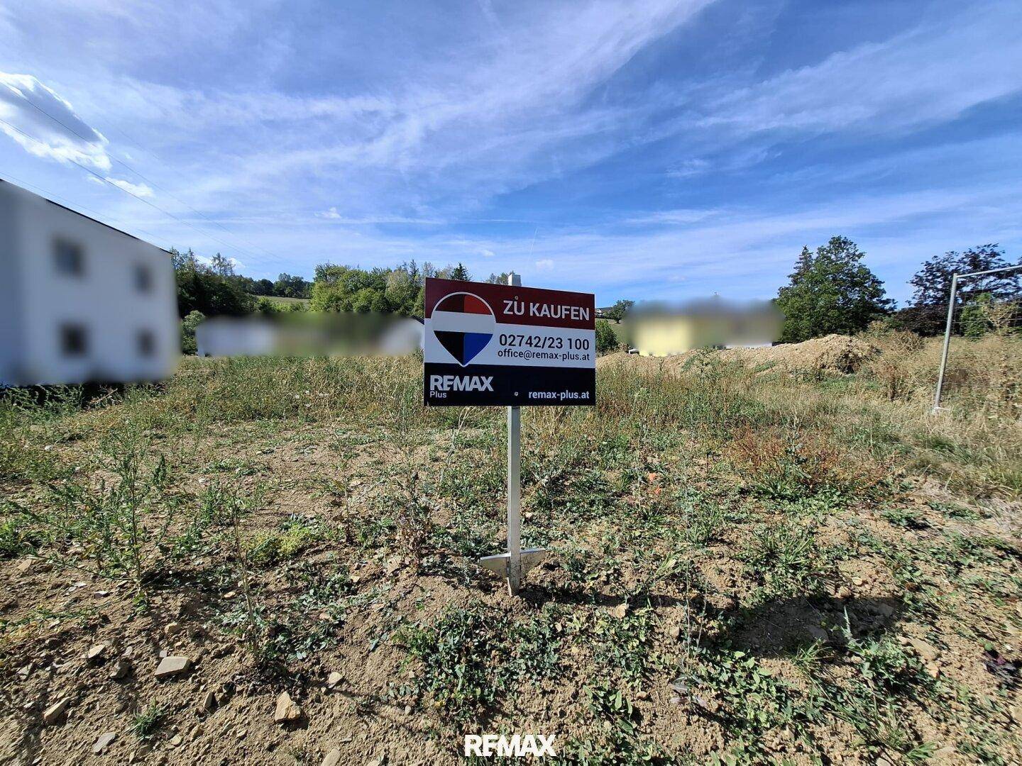 Grundstück mit einem 'Zu Kaufen'-Schild, umgeben von Gras und Bäumen unter blauem Himmel.